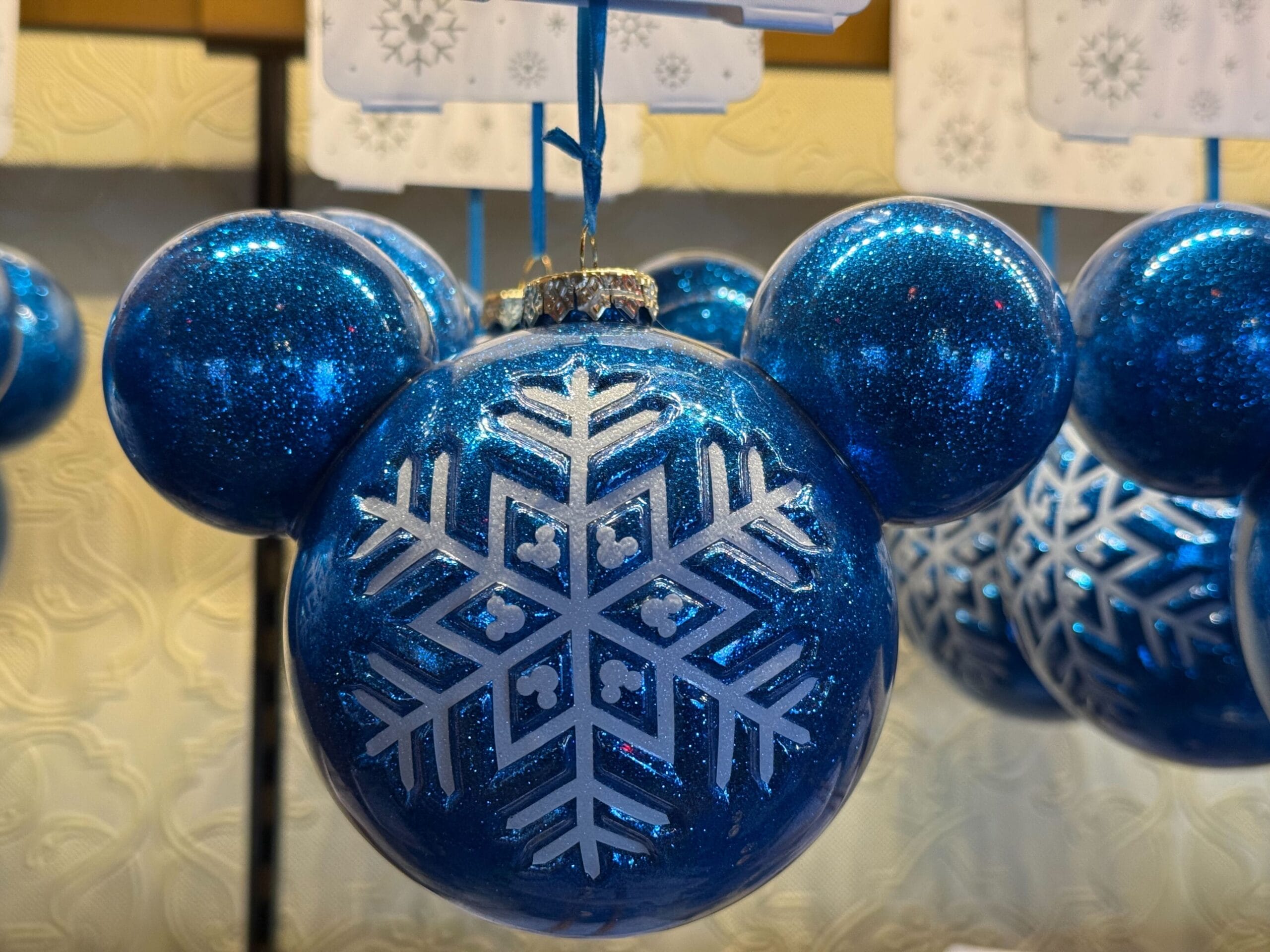Blue Mickey Mouse-shaped ornament with snowflake motif among 2025 Disneyland holiday merchandise; Sleeping Beauty Castle seen nearby.