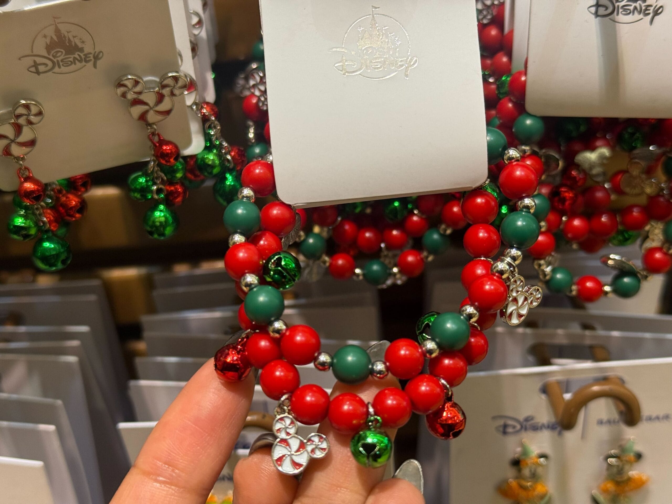Hand holding a Disneyland holiday bracelet with red and green beads, jingle bells, and park-exclusive Christmas merchandise on display.