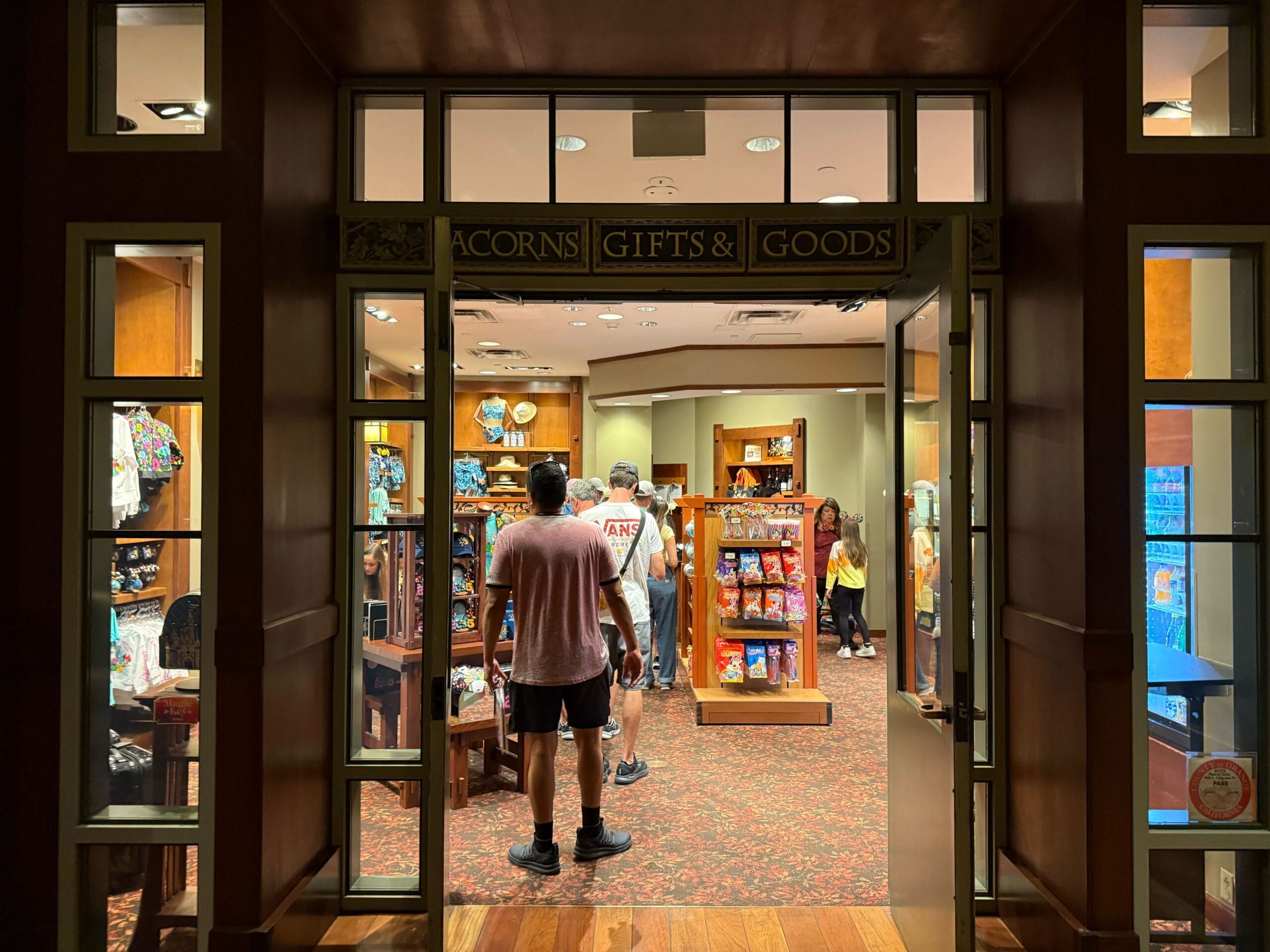 Guests explore shelves filled with Mickey merchandise inside Acorns Gifts & Goods, a cozy Disney park shop aglow with warm lights.