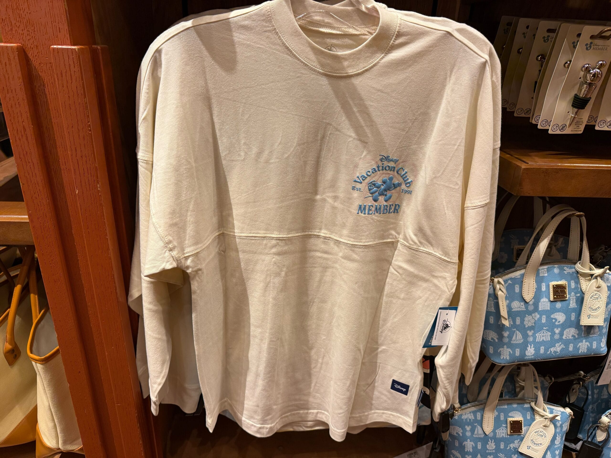 A white "Vacation Club Member" long-sleeve shirt hangs by blue bags—classic Disney Parks merch for any Disneyland or WDW fan.