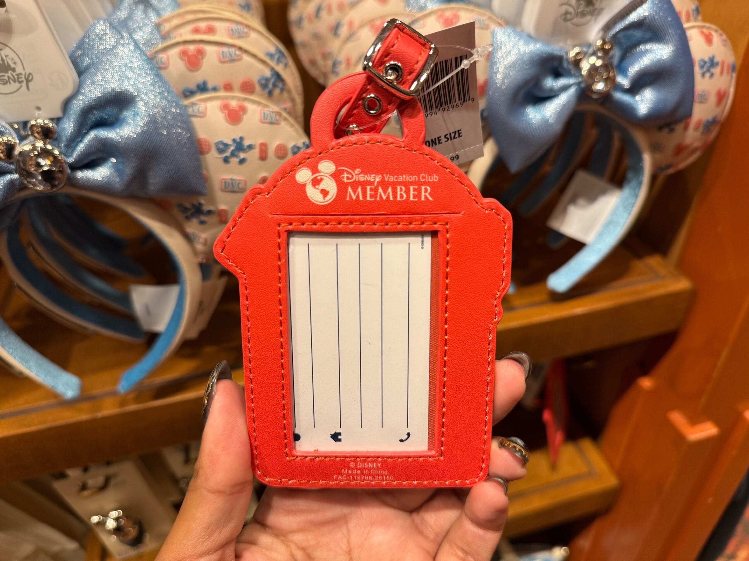 A hand displays a red DVC Member tag by Mickey merch and Minnie ear headbands inside a Disneyland shop, not far from Sleeping Beauty Castle.