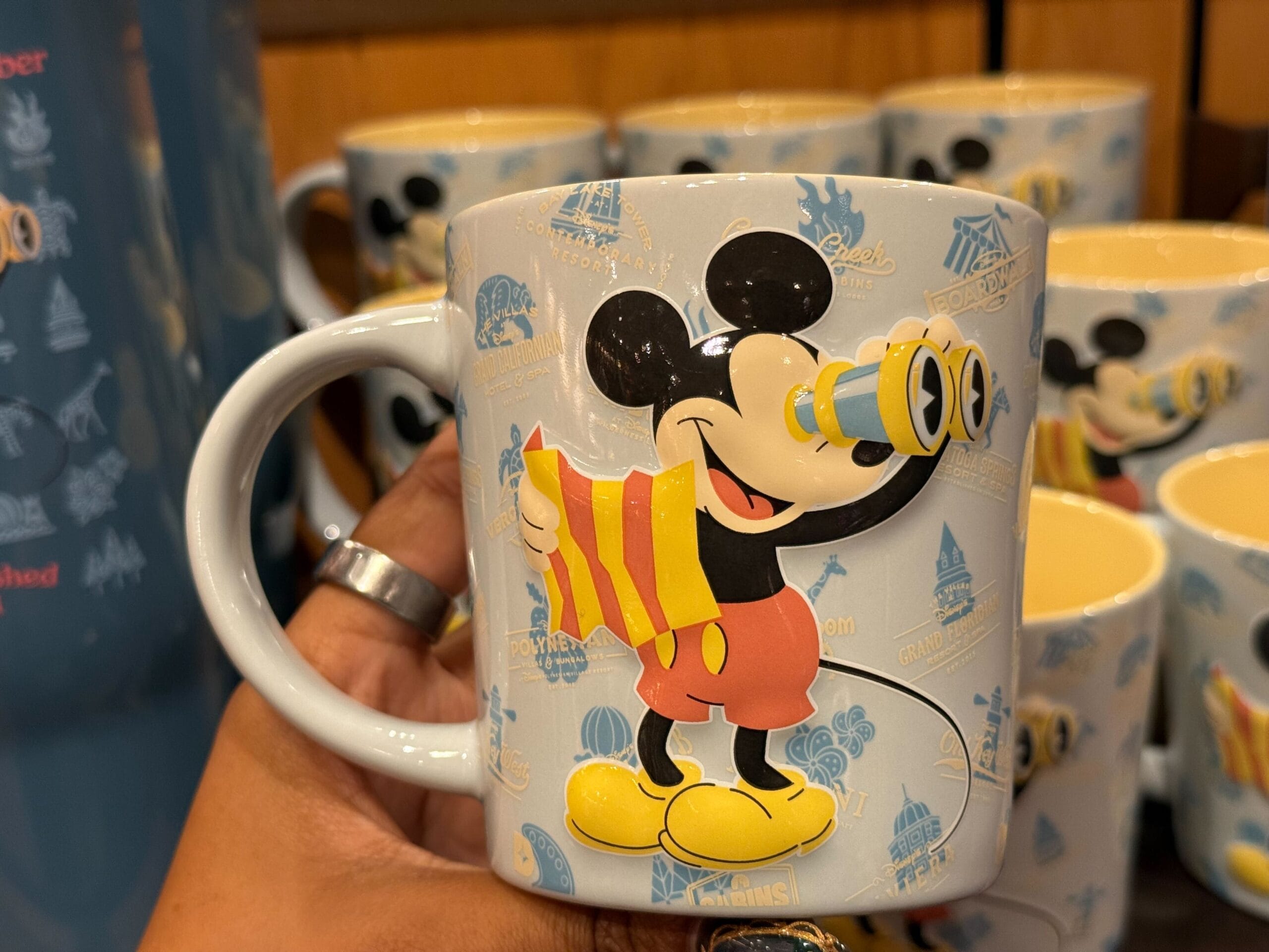 A hand holds a Mickey Merch mug with Mickey Mouse and binoculars, ideal for DVC Members; extra mugs line up like at Disney gift shops.