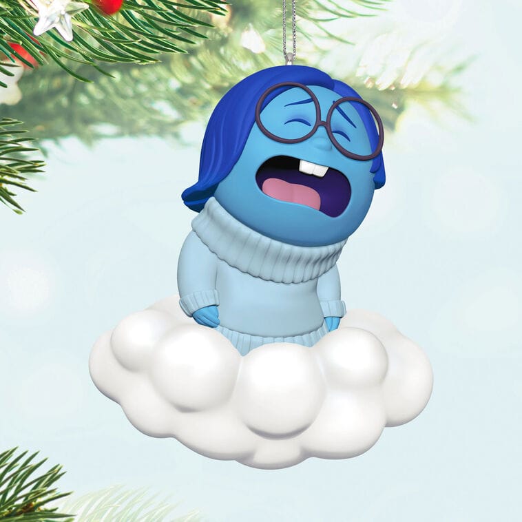 A blue cartoon character with glasses sits sadly on a Hallmark ornament cloud, hanging on a Christmas tree like Disney park decor.
