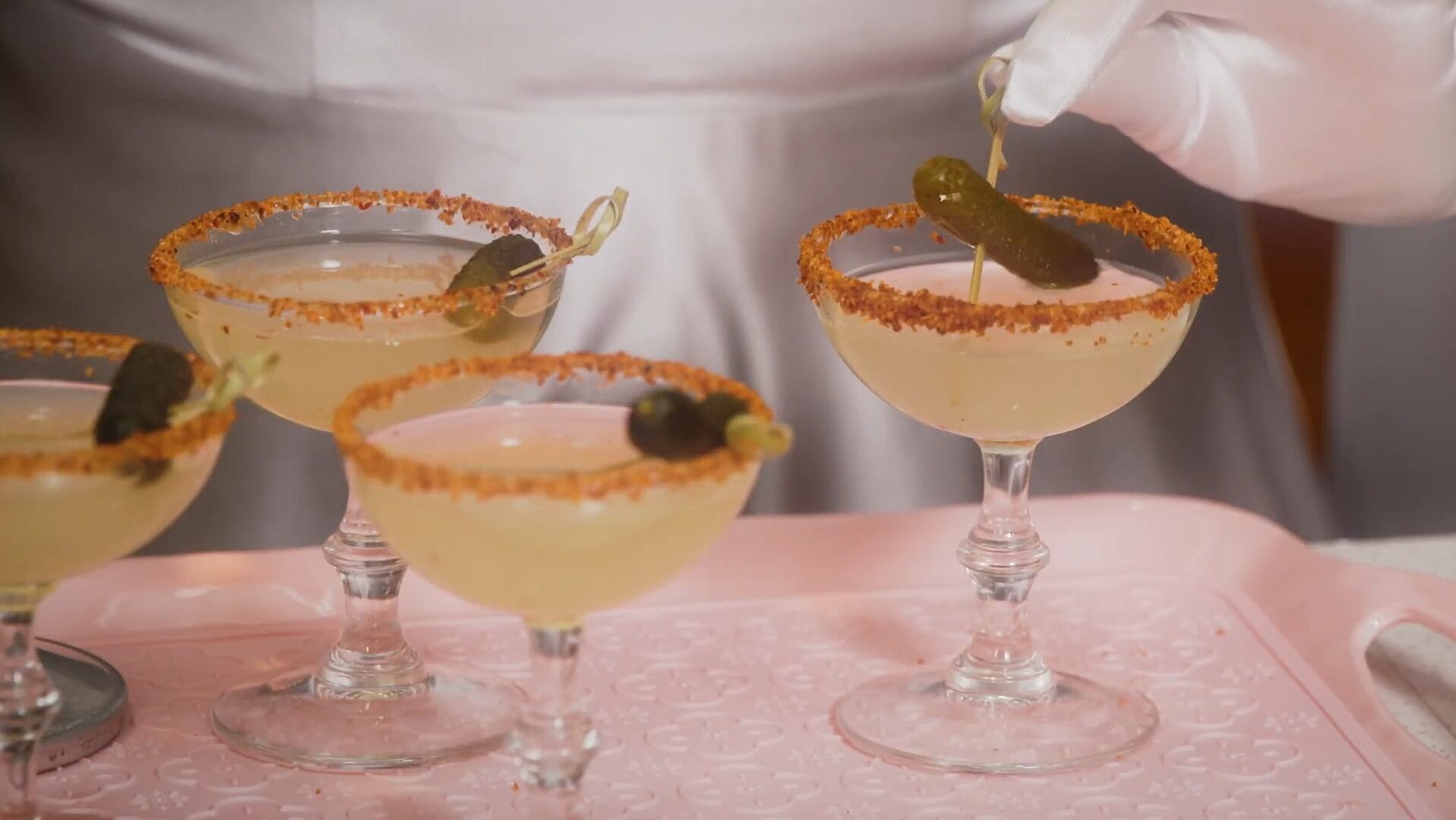 Three spicy cocktails with chili-rimmed glasses and pickles on a pink tray at Disney Jollywood Nights, no castle pictured.