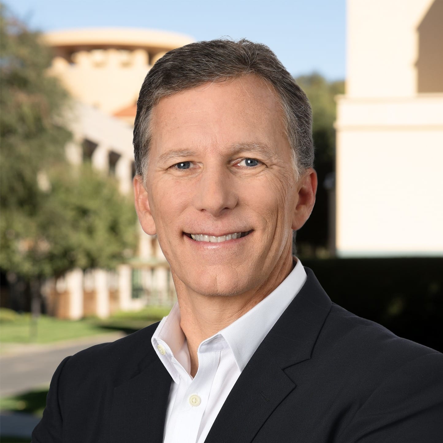 Smiling man in a suit stands outside with Disney park-style buildings and trees behind him, but no iconic castle visible.