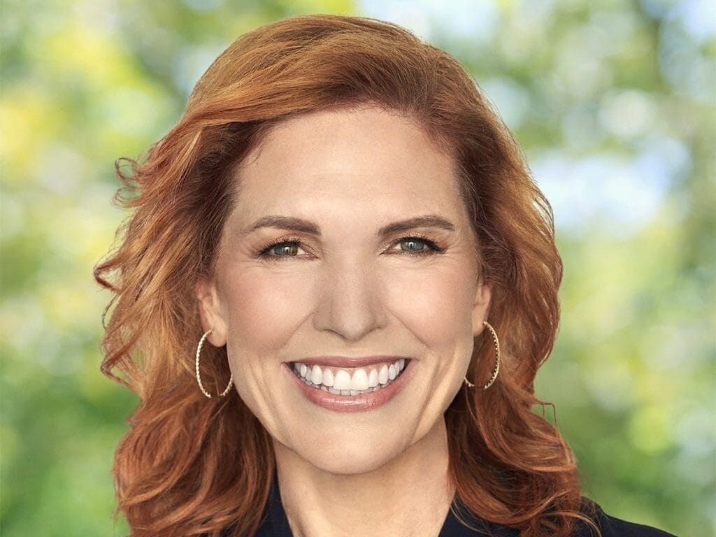 Smiling woman with wavy red hair and hoop earrings, wearing a dark top, standing outside at what could be a Disney park location.