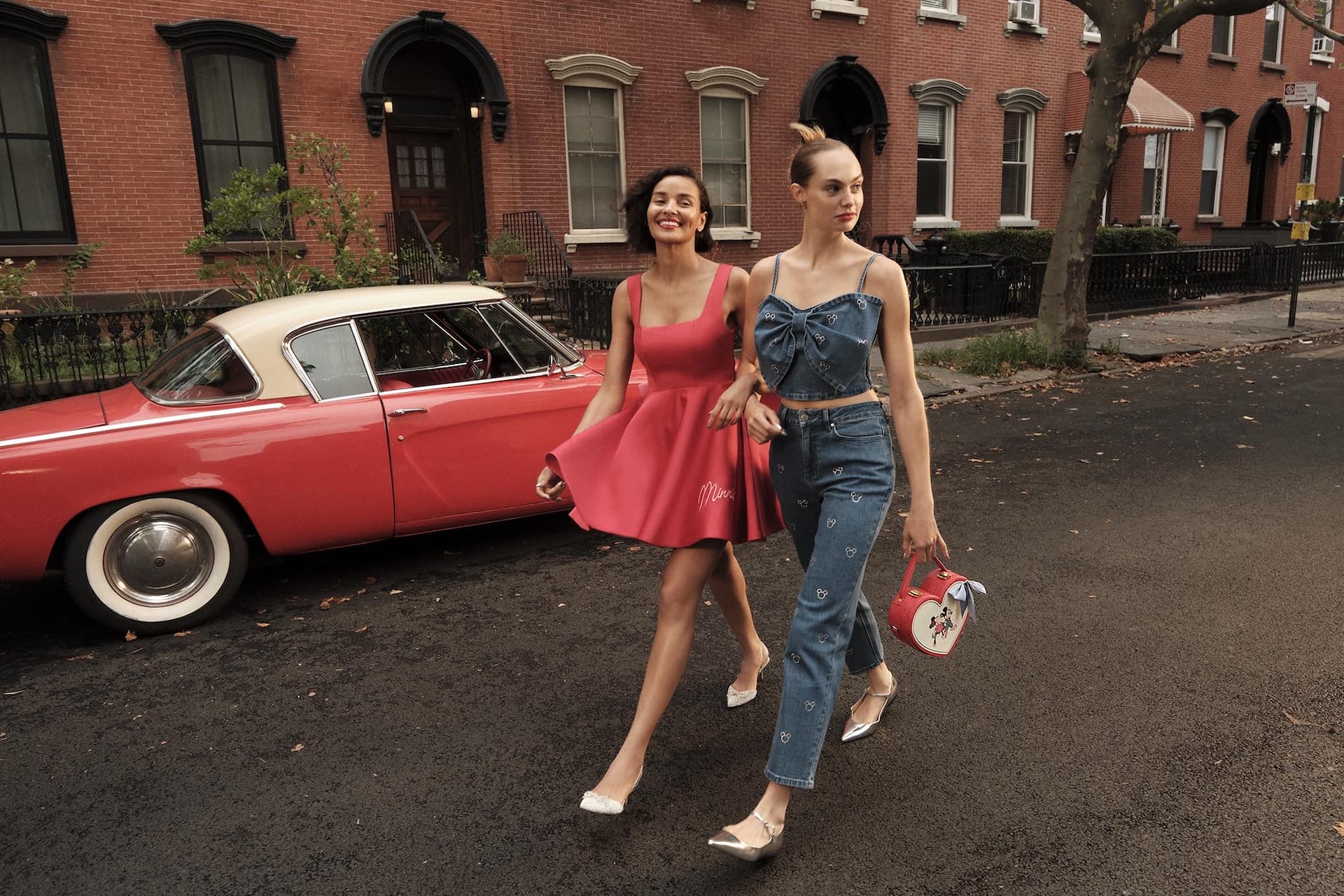 Two women walk by a vintage red car on a city street, one in denim, the other in a Macy's Holiday pink dress—no Disney castles seen.