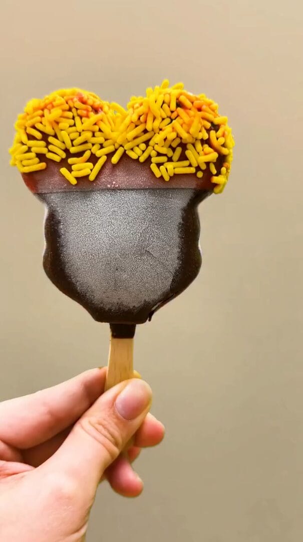 A hand holds a Mickey's Premium Ice Cream bar with the top half dipped in milk chocolate with yellow sprinkles.