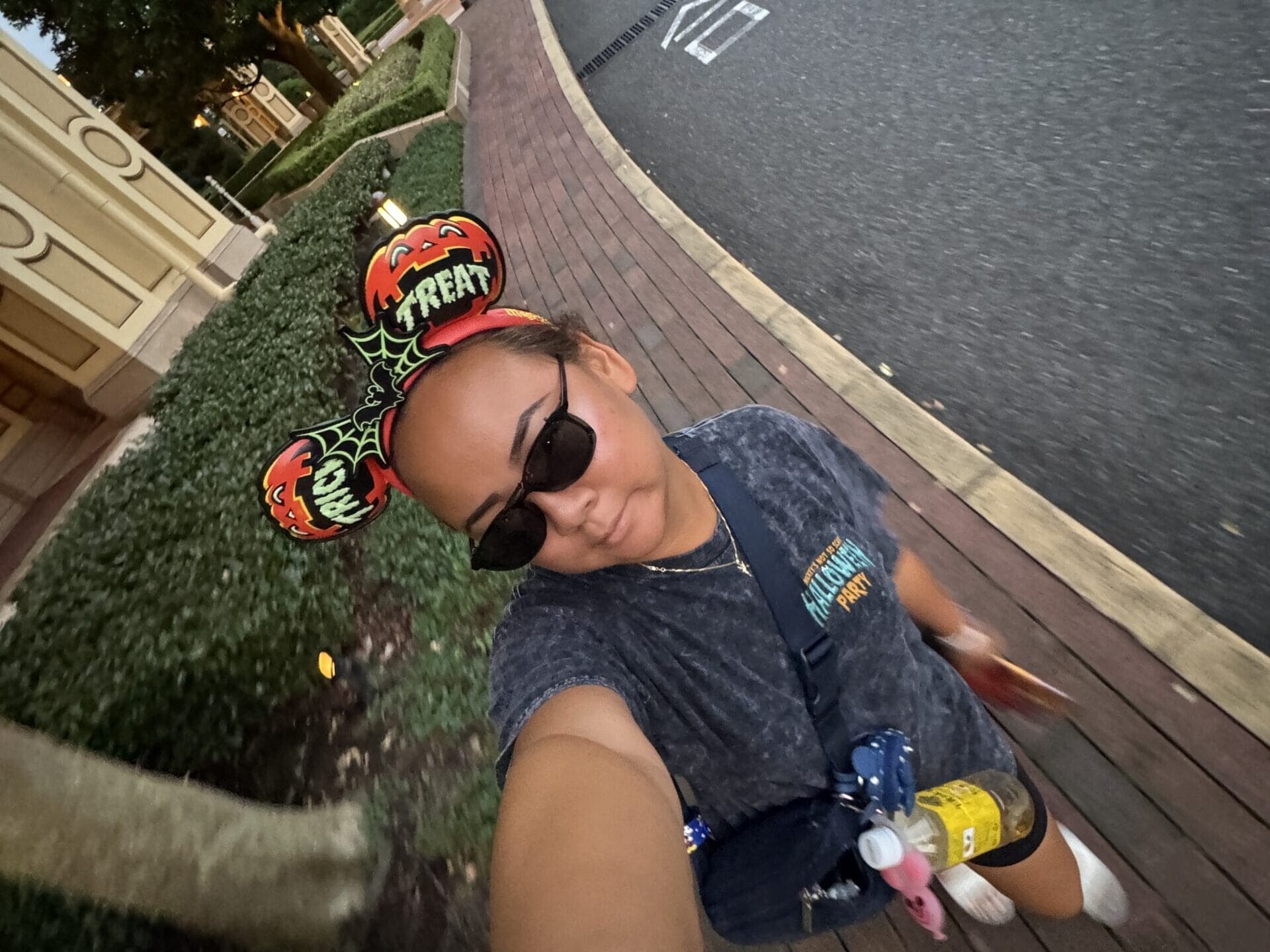 Guest in Halloween-themed Mickey ears and sunglasses poses for a selfie at Tokyo Disney Resort, with trees and buildings behind.