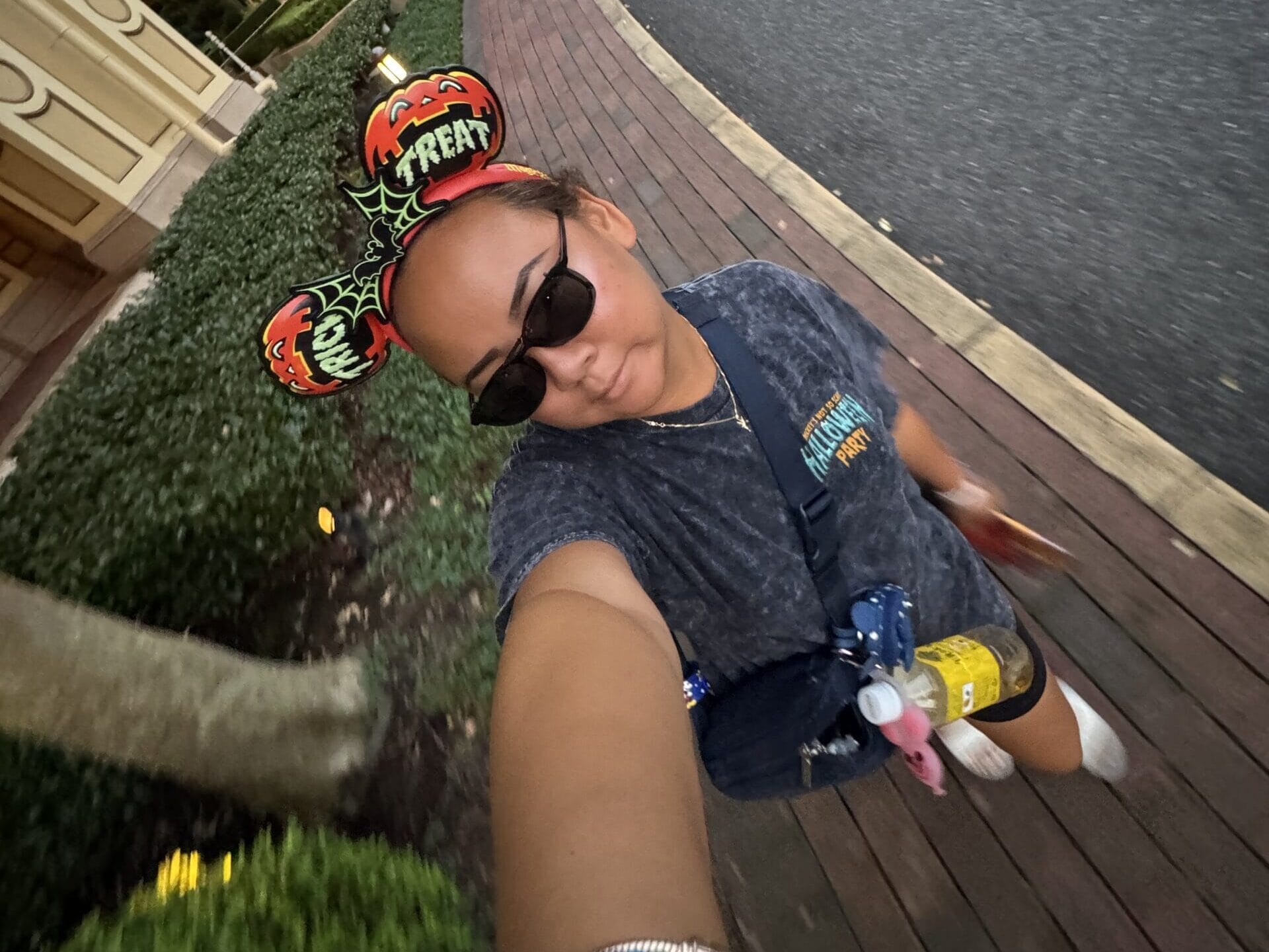 Guest in Halloween-themed Mickey ears and sunglasses poses for a selfie at Tokyo Disney Resort, with trees and buildings behind.