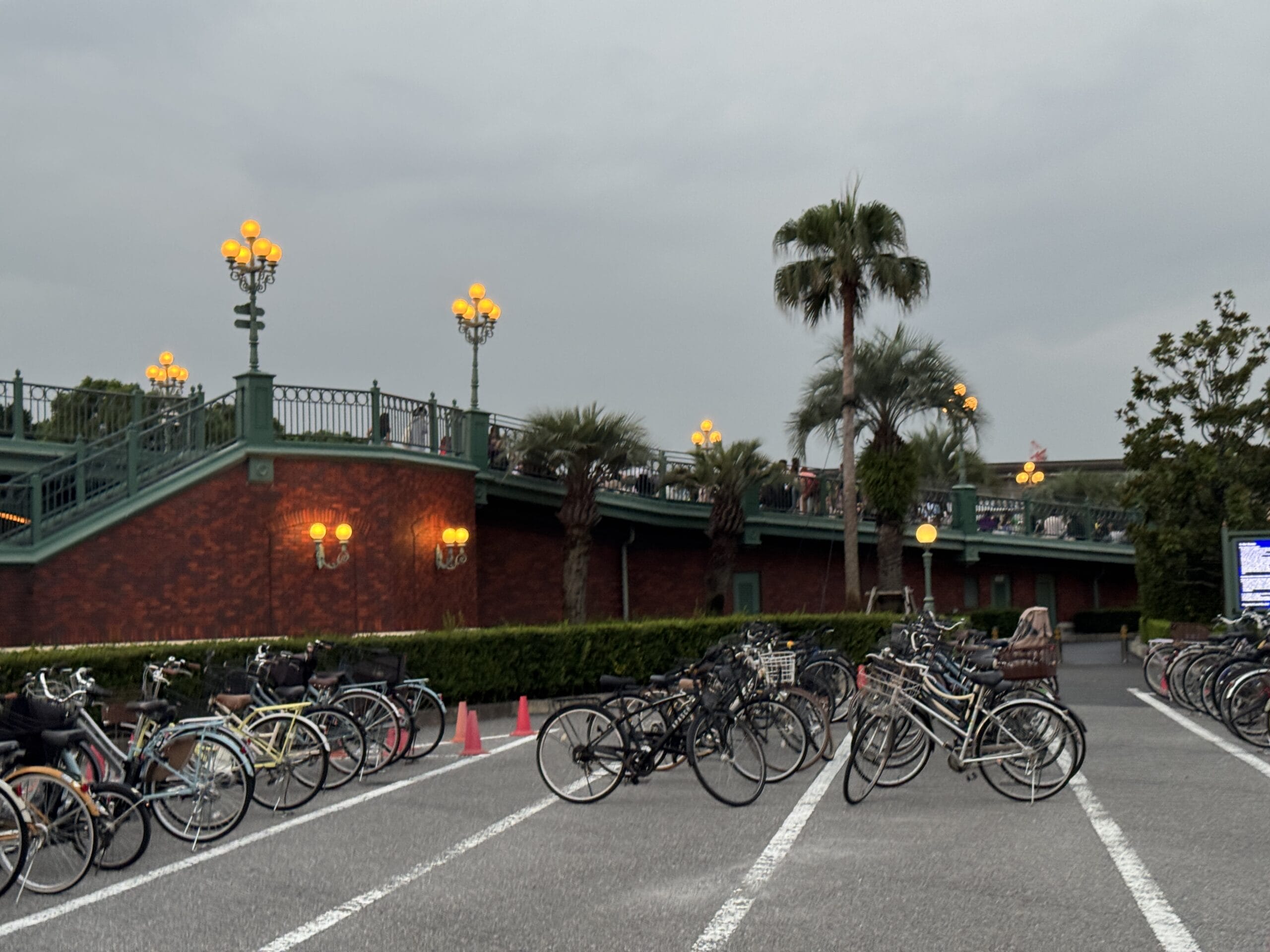 Bicycles by a brick bridge, glowing street lamps, and palm trees, capturing Tokyo Disney Resort’s atmosphere on a cloudy day.