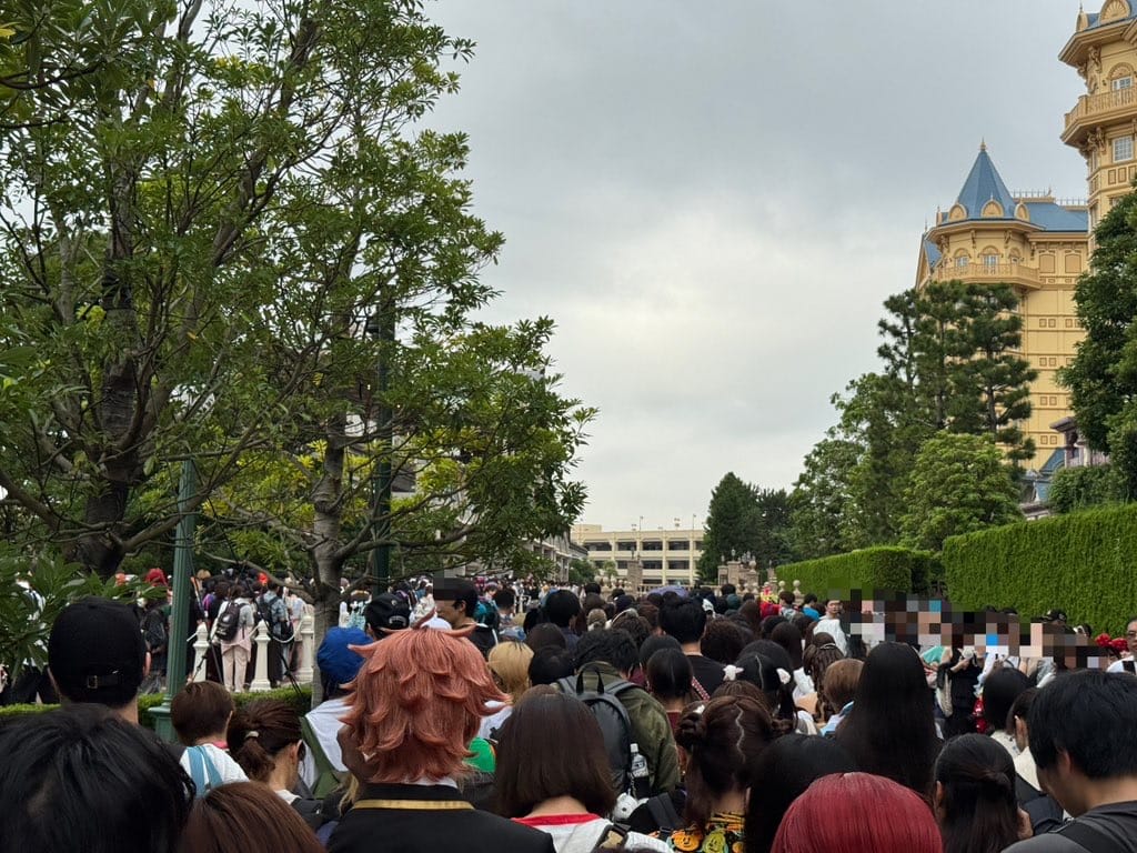 A large crowd, some in Halloween costumes, gathers by trees and a tall building that doesn’t resemble any Disney castle, under clouds.