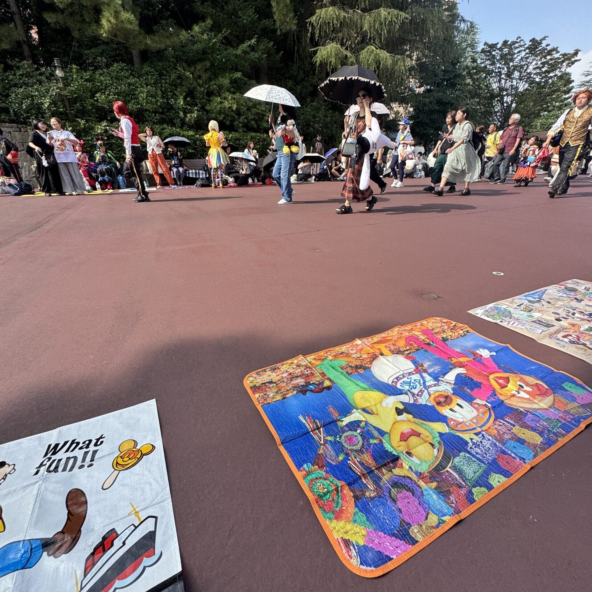 Guests in costumes stroll on the red pavement at Tokyo Disney Resort, with vibrant blankets and art displayed, no castle in view.