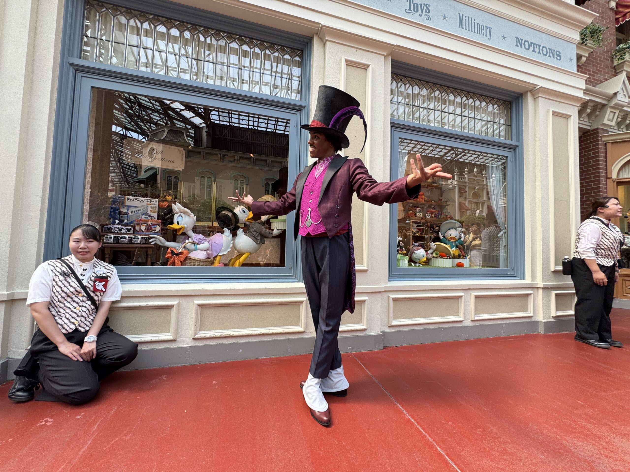 A performer in a tall hat and purple coat stands by a Tokyo Disney Resort toy store, plush toys behind—no castle like Disneyland’s.
