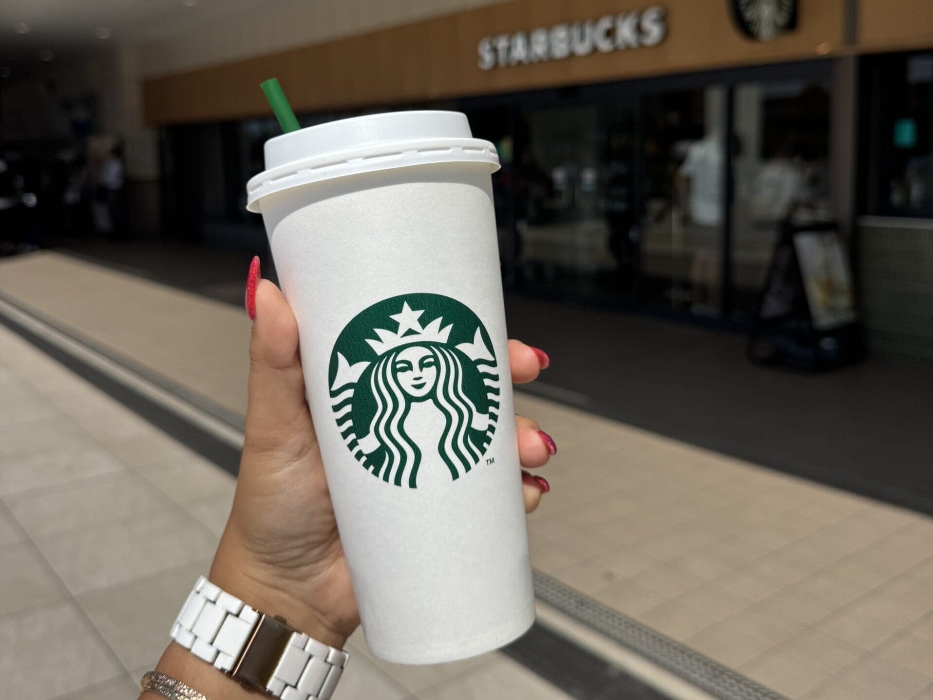 A hand holds a Starbucks cup outside the Starbucks at Tokyo Disney Resort, with Cinderella Castle nearby on a bright sunny day.
