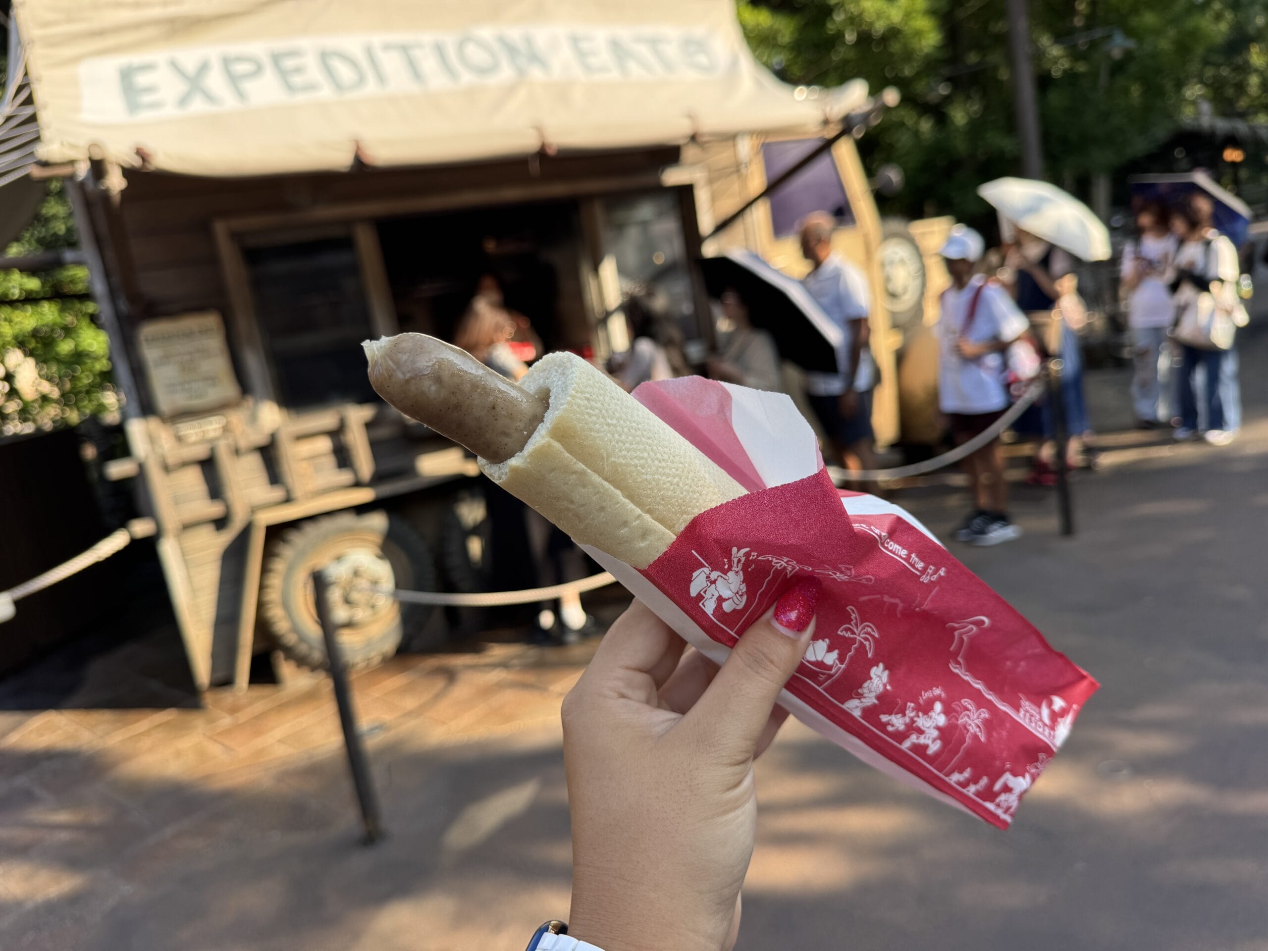 A sausage in a bun is held up near Expedition Eats at Disney's Hollywood Studios, with park guests visible in the background.