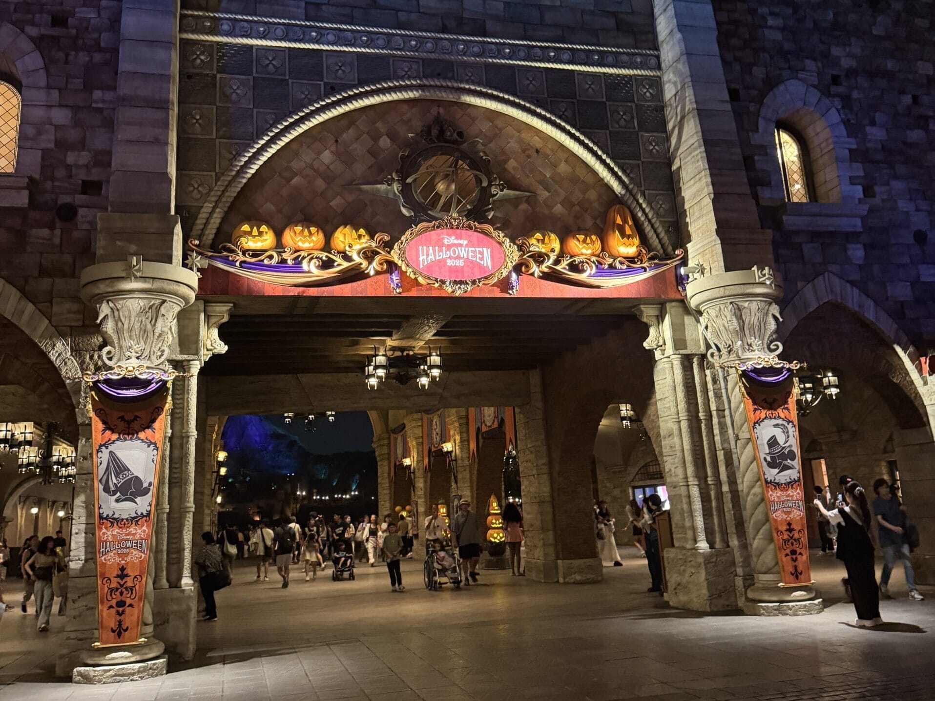 Glowing jack-o'-lanterns and Halloween banners decorate Tokyo Disney Resort's arched entrance as guests walk beneath at night.