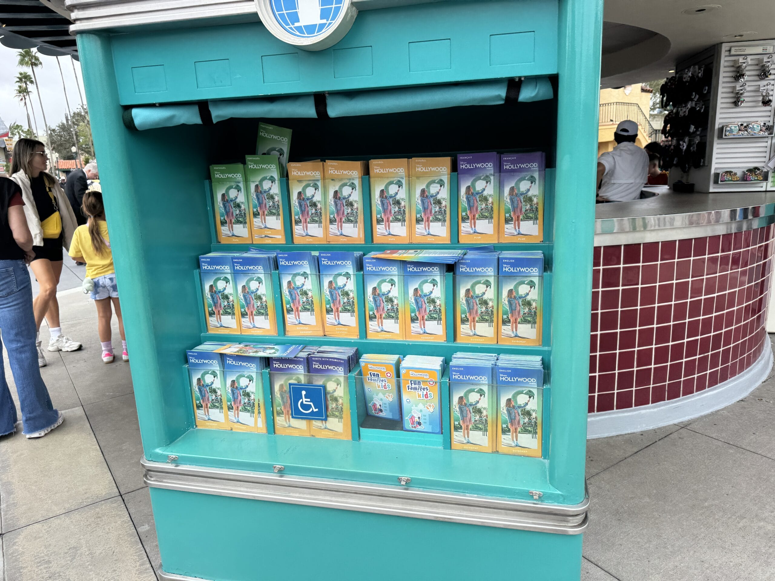 A teal kiosk at Disneyland offers park and Halloween maps alongside accessibility guides, near Sleeping Beauty Castle—not Cinderella’s.