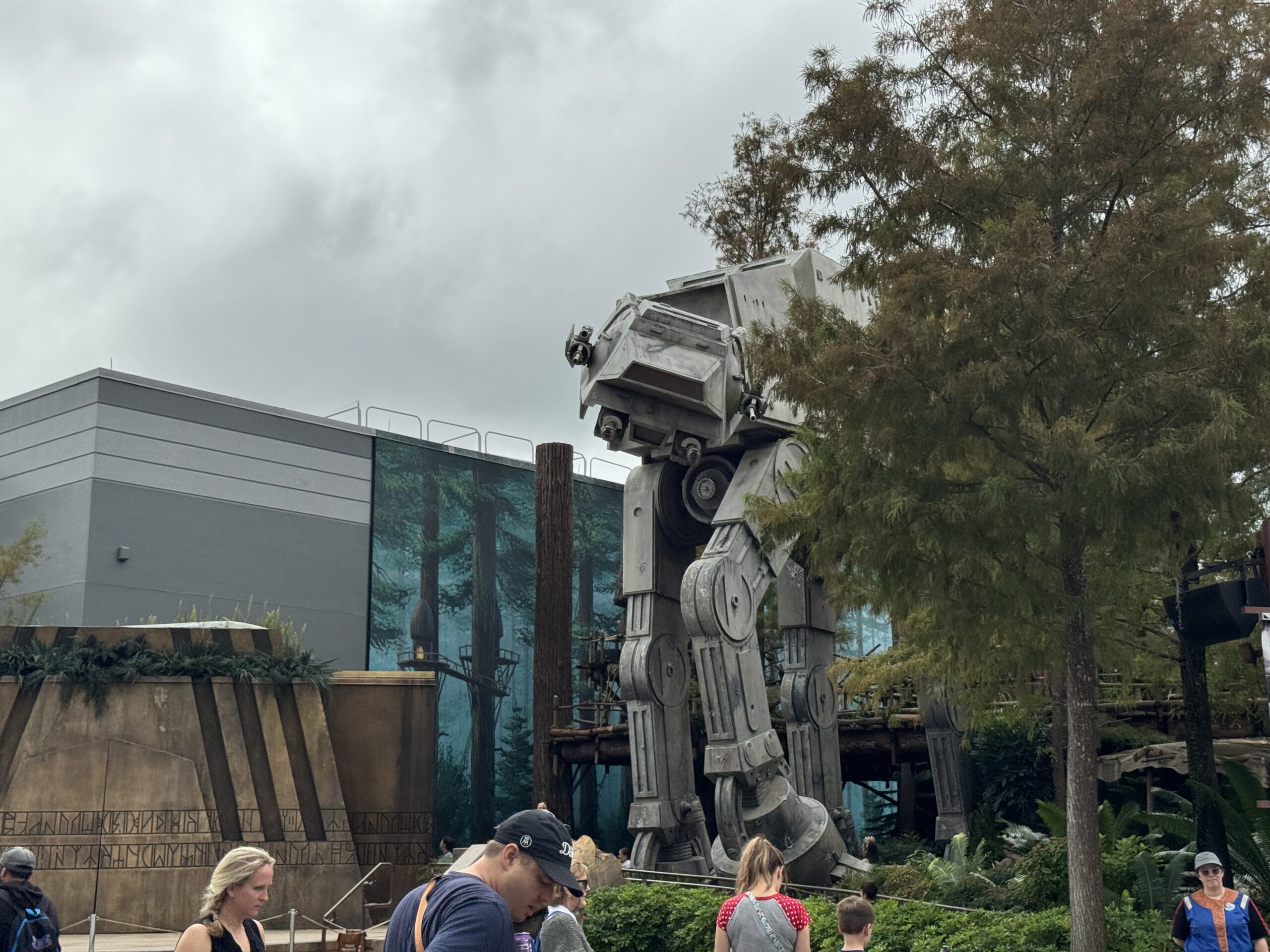 Visitors walk past a life-size AT-AT walker at Disney's Hollywood Studios, surrounded by trees and a building in the park.
