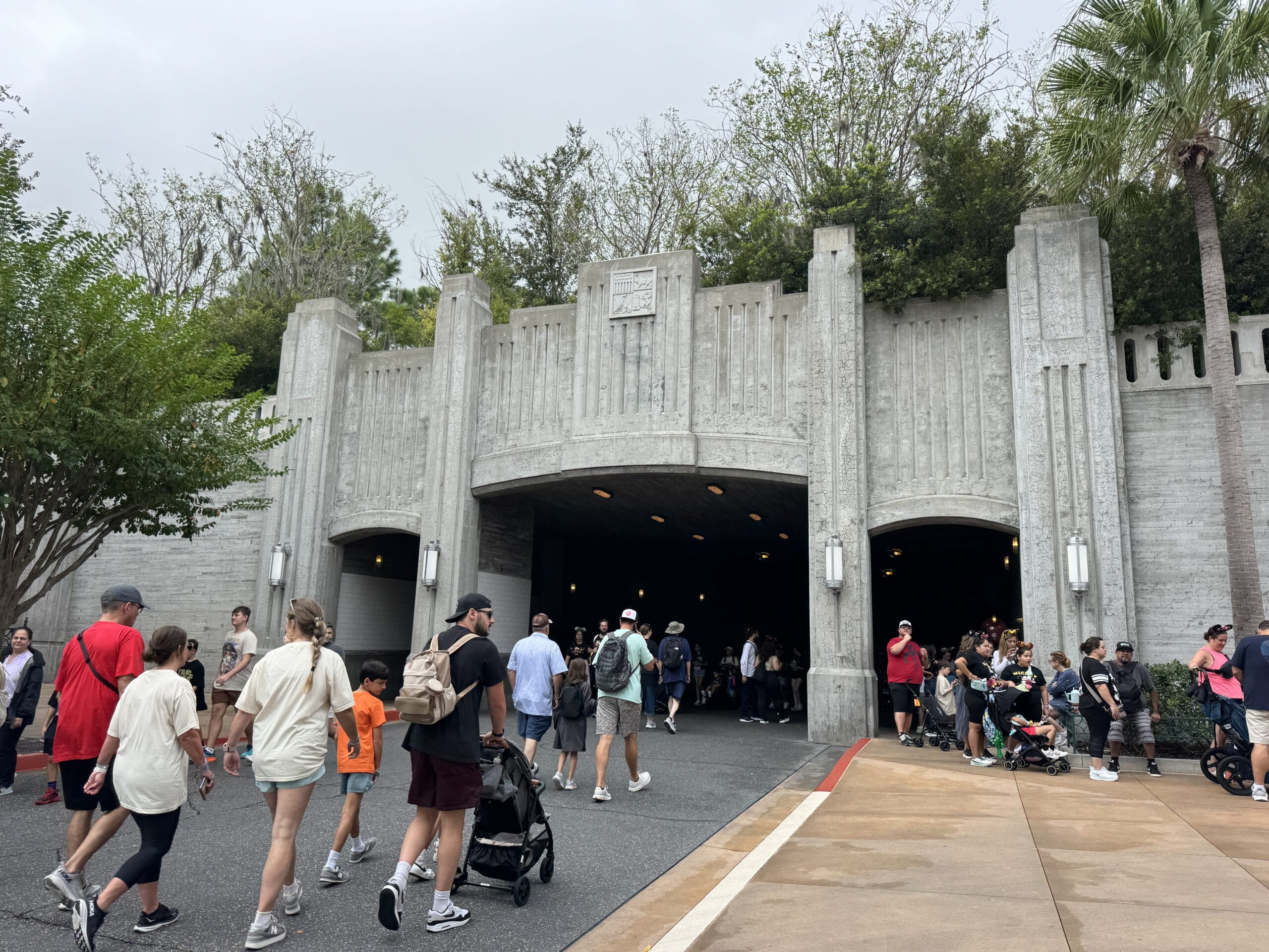 Visitors stroll by a grand, gray Art Deco tunnel entrance at Tokyo Disney Resort, which lacks the iconic fairy-tale castle seen in other Disney parks like Cinderella Castle in Walt Disney World or Sleeping Beauty Castle at Disneyland, all under overcast skies.