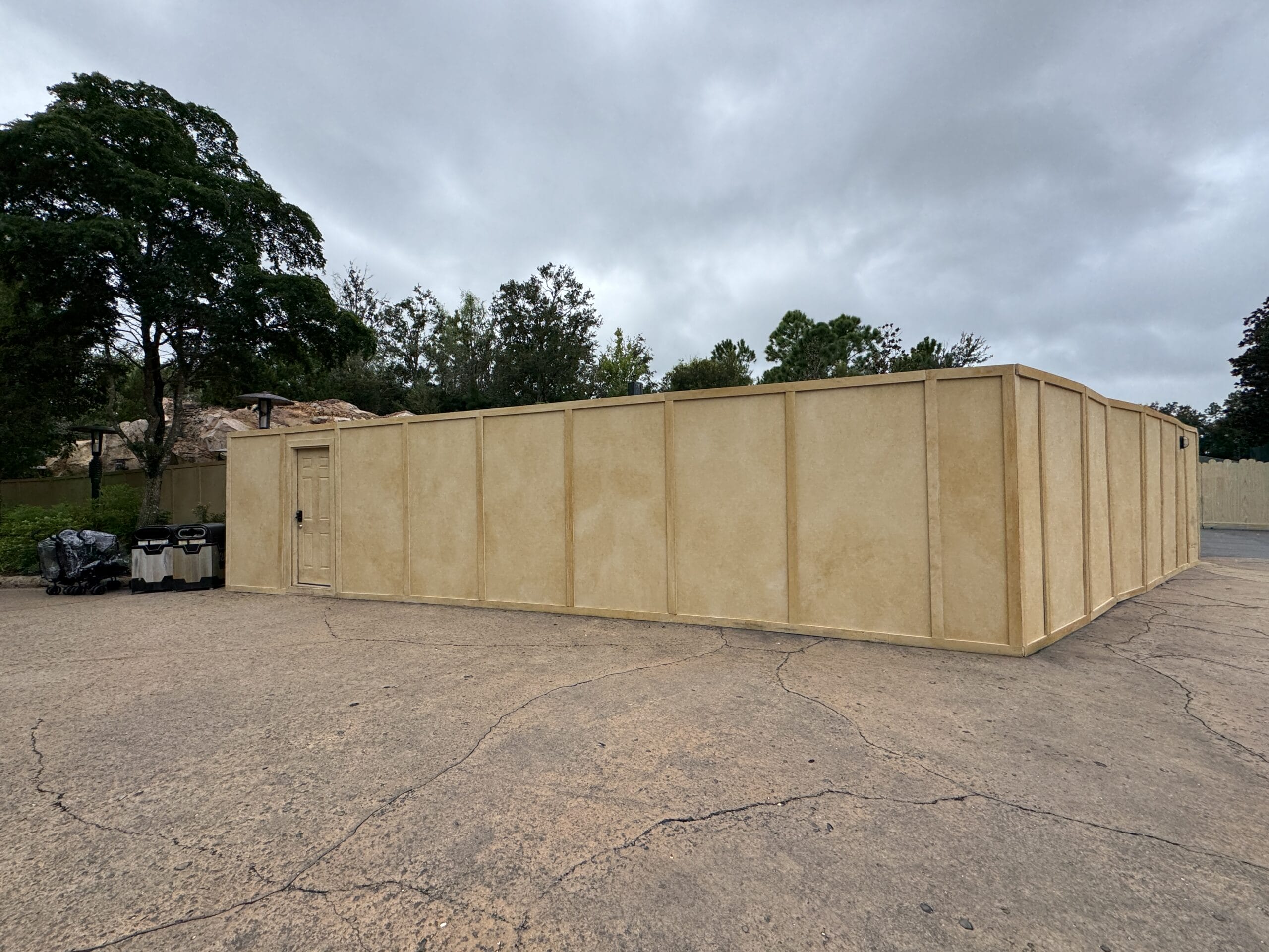 Tan construction walls at Disney's Hollywood Studios enclose an area, with trees and a cloudy sky—no castle like the Magic Kingdom's.