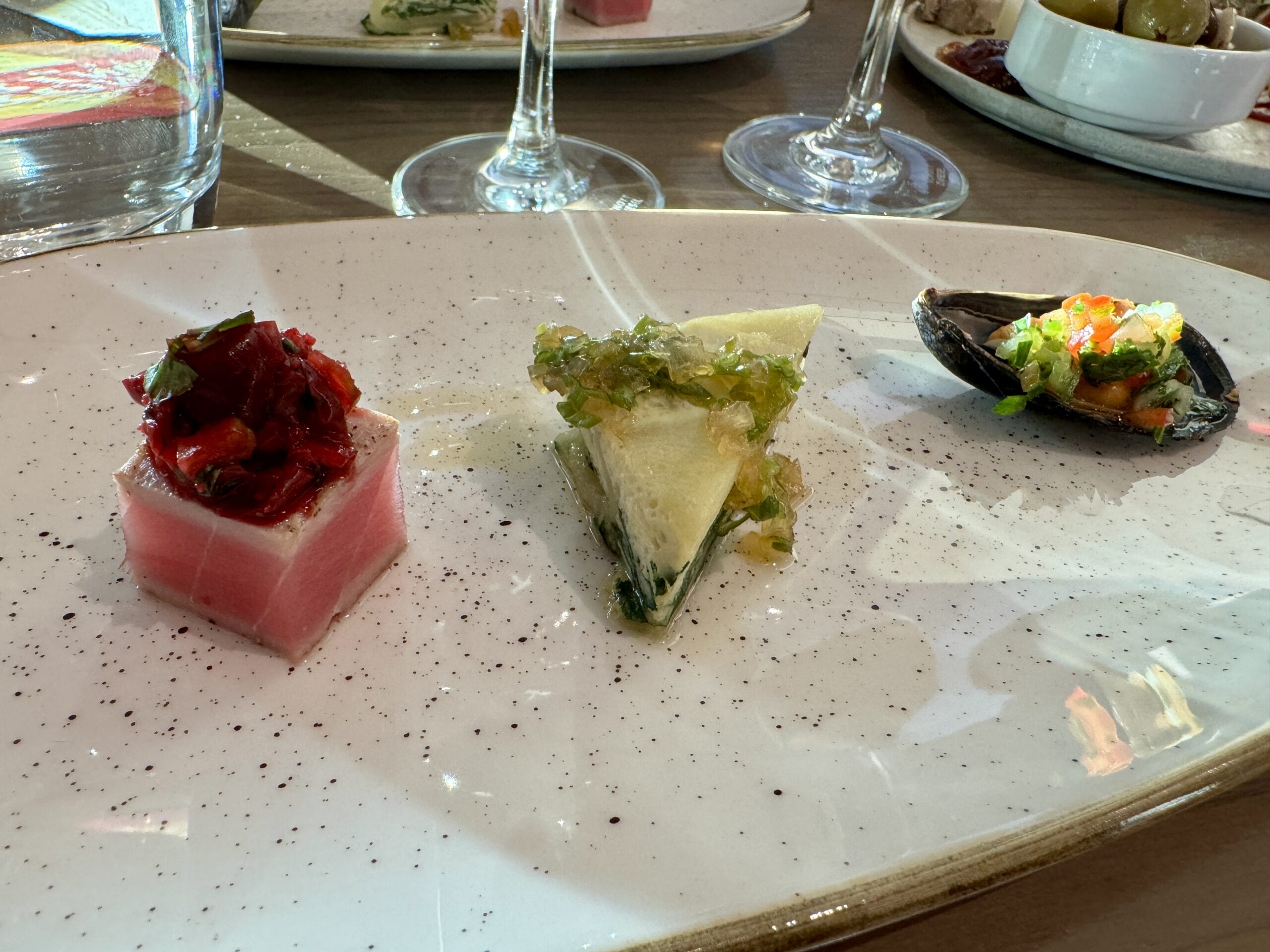 Three small gourmet tapas are artistically arranged on a white plate with wine glasses in the background.