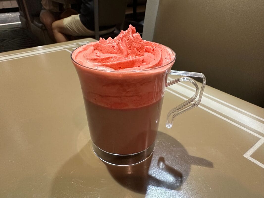 A glass mug of hot chocolate with pink whipped cream sits on a tan table, themed for Jollywood Nights at a Disney park.