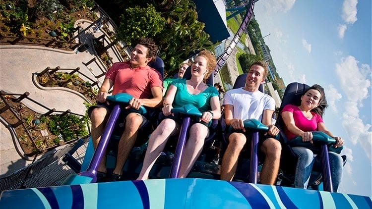 Four riders laugh and smile on a steep Mako Coaster drop, with a theme park view below—.