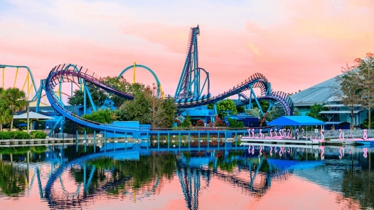 A blue roller coaster winds above a serene lake at sunset, its reflections reminiscent of Epcot's vibrant evenings in Disney Parks.