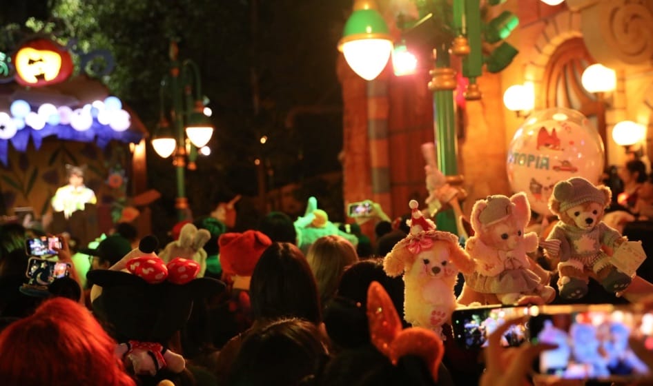 Disney park guests at night pose with plushies and snap photos by vibrant lights; no classic Disney castle visible in scene.