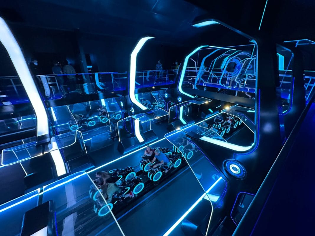TRON: Ares-themed indoor roller coaster station at a Disney park, aglow with blue lights and guests boarding light cycles.