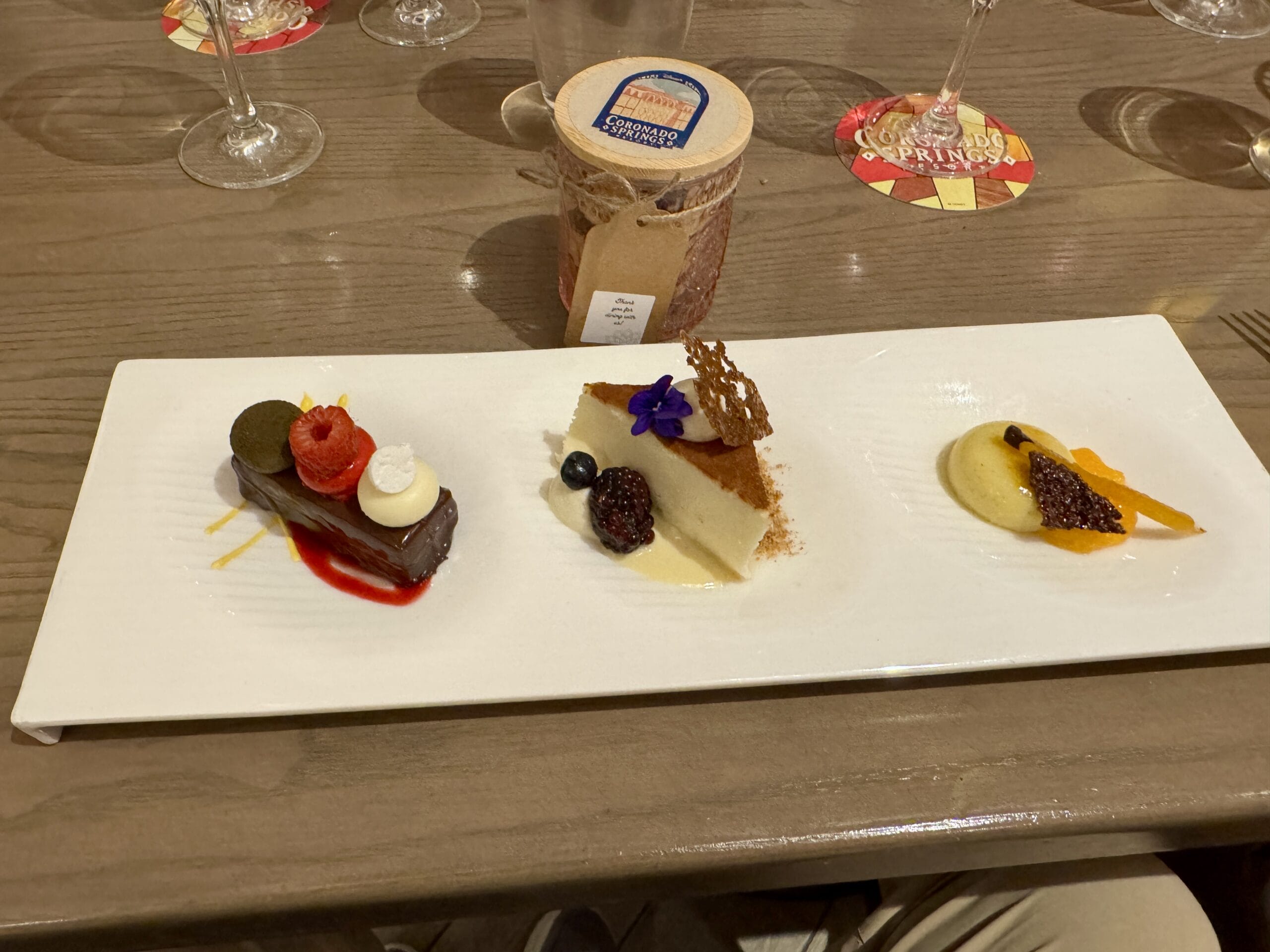 White plate with three gourmet desserts, berries, and garnishes; perfect for a Toledo tapas night with wine glasses in the background.