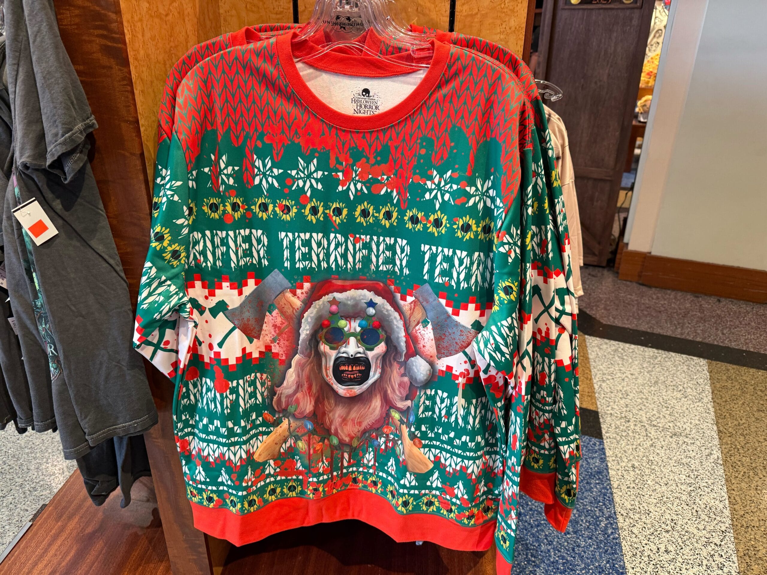 Ugly Christmas sweater featuring Art the Clown with Christmas tree glasses and a Santa hat in front of crossed bloody axes. Faux blood splatter drips down from the shoulders.