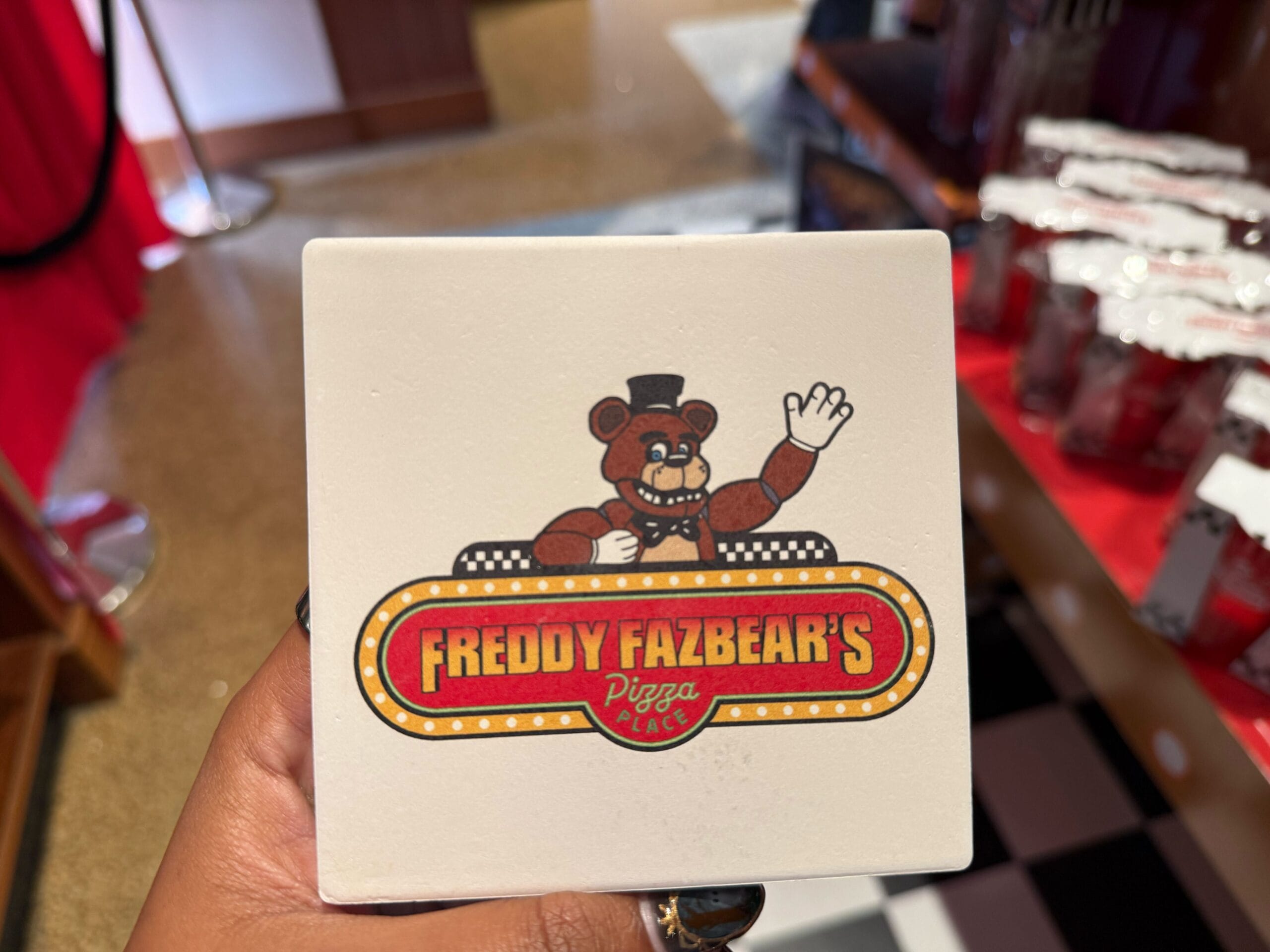 A hand holds a square coaster featuring the Freddy Fazbear's Pizza Place logo.