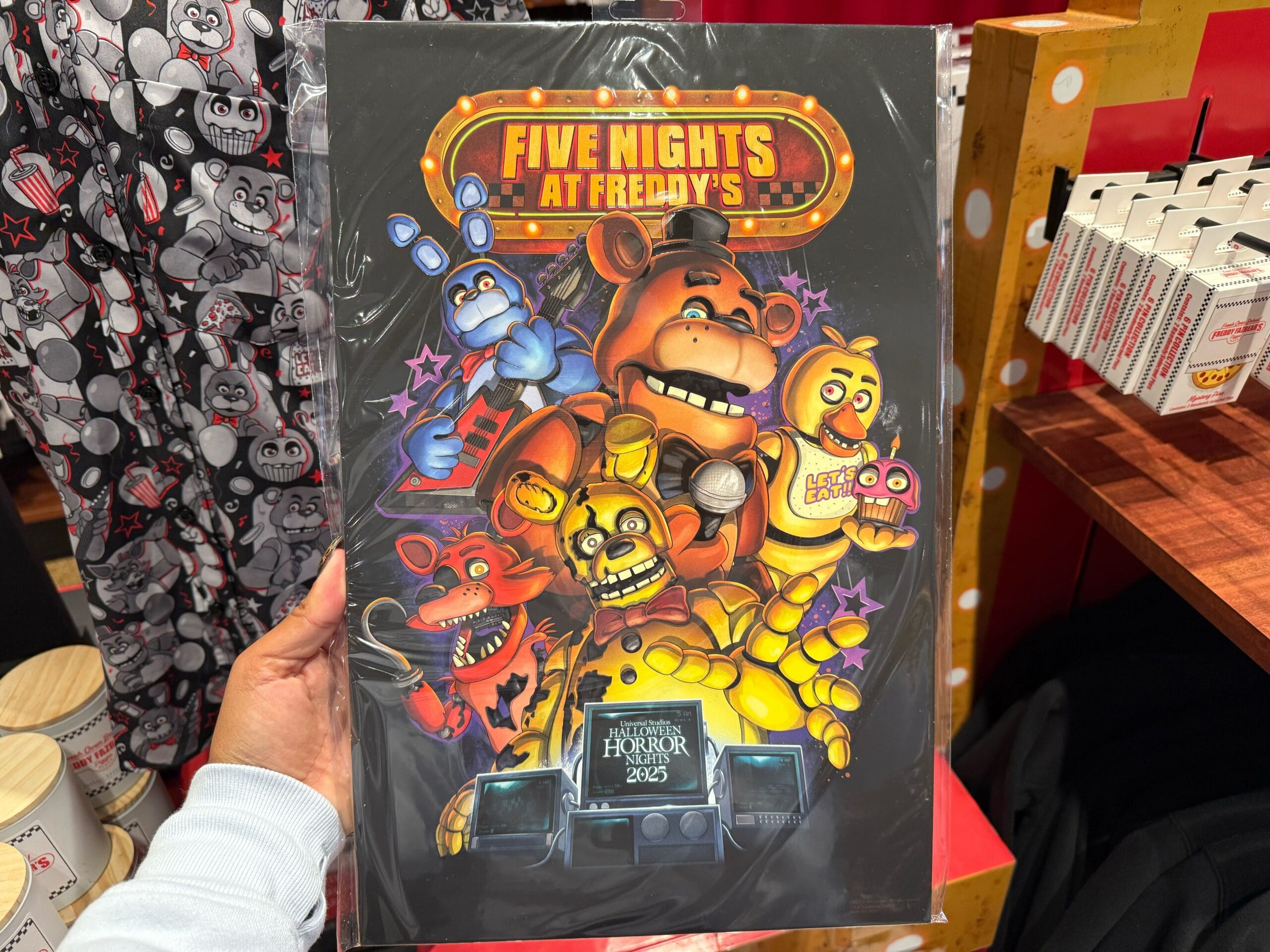 A vibrant "Five Nights at Freddy's" poster featuring the five animatronics--Bonnie, Foxy, Springtrap, Freddy, and Chica with Cupcake--above security monitors displaying "Universal Studios Halloween Horror Nights 2025."
