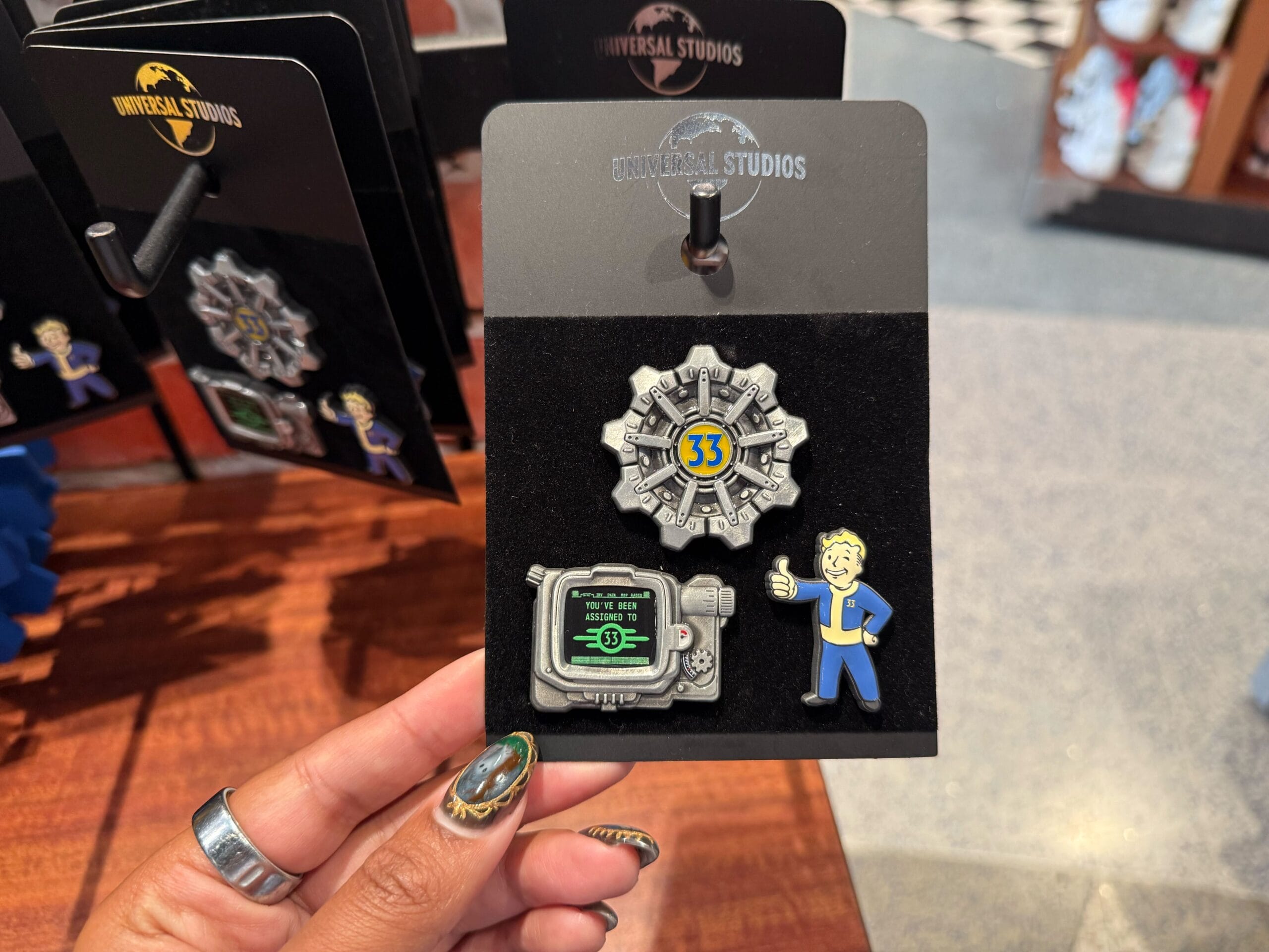 A hand holds a Universal Studios Fallout pin set with Vault 33 door, Vault Boy, and Pip Boy pins.