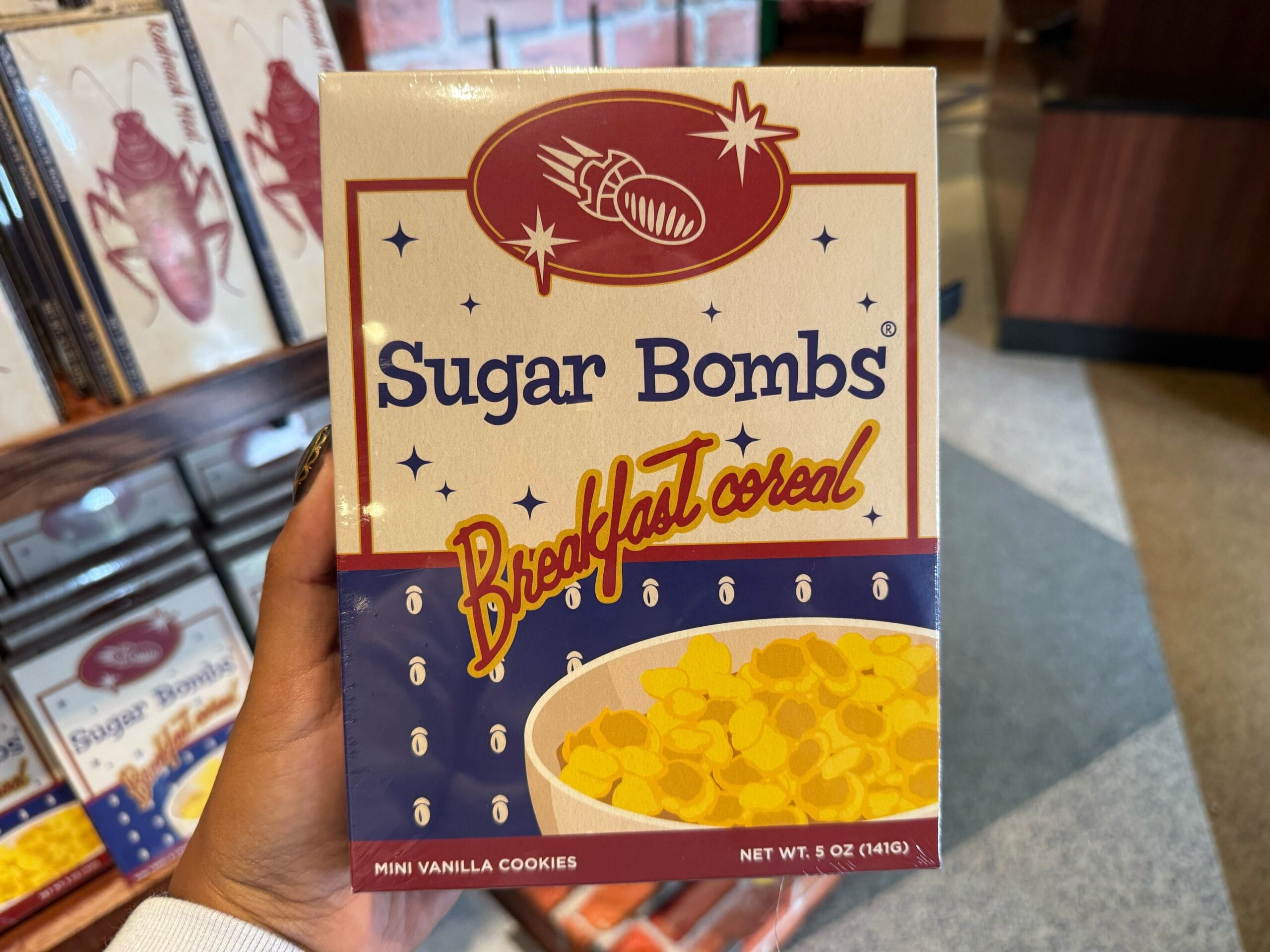 A hand holds a box of Sugar Bombs breakfast cereal, unique Fallout merch from Universal Studios.