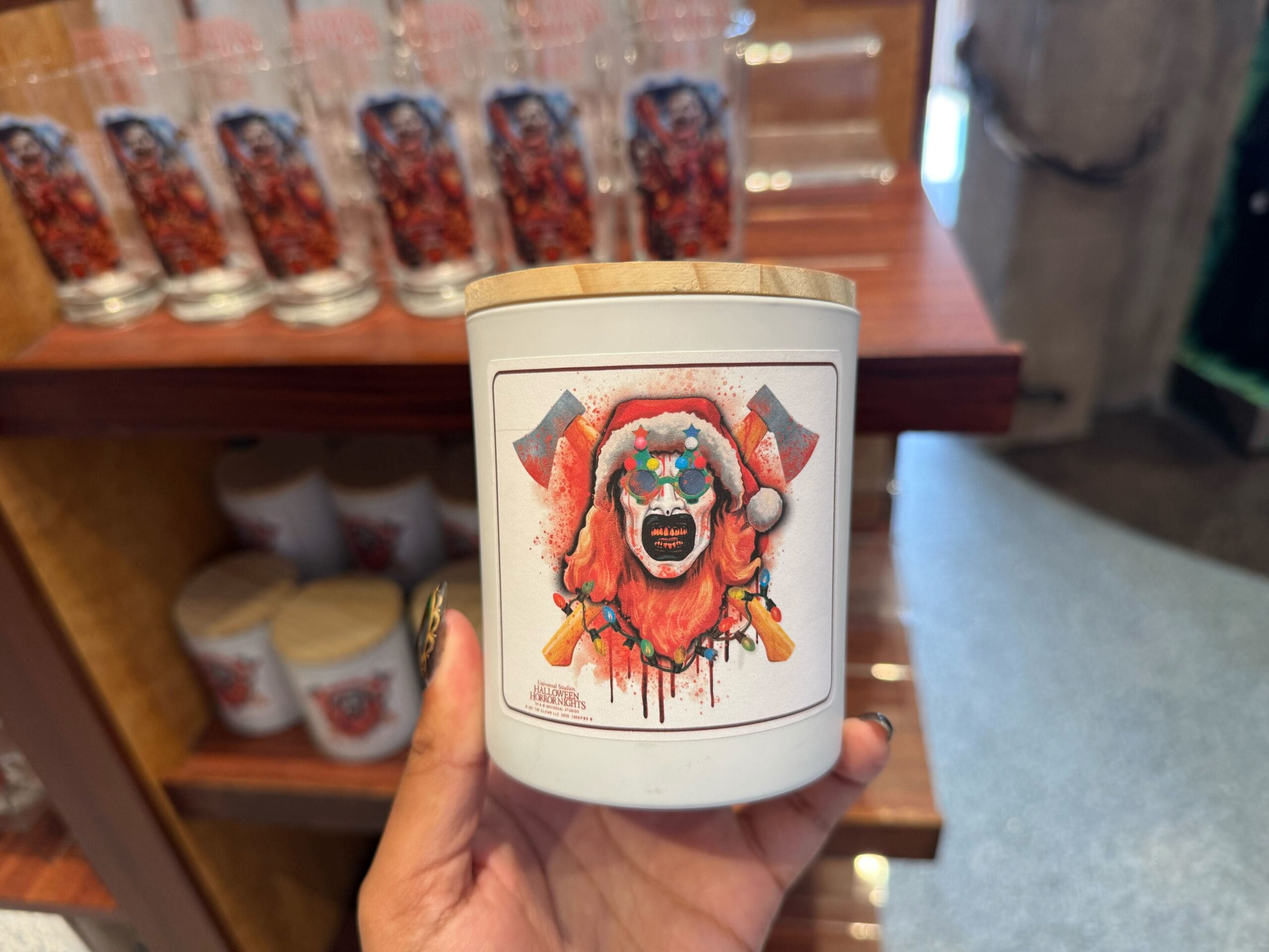 A 2025 Halloween Horror Nights lidded candle jar with Art the Clown in Christmas tree glasses and a Santa hat in front of bloody crossed axes.