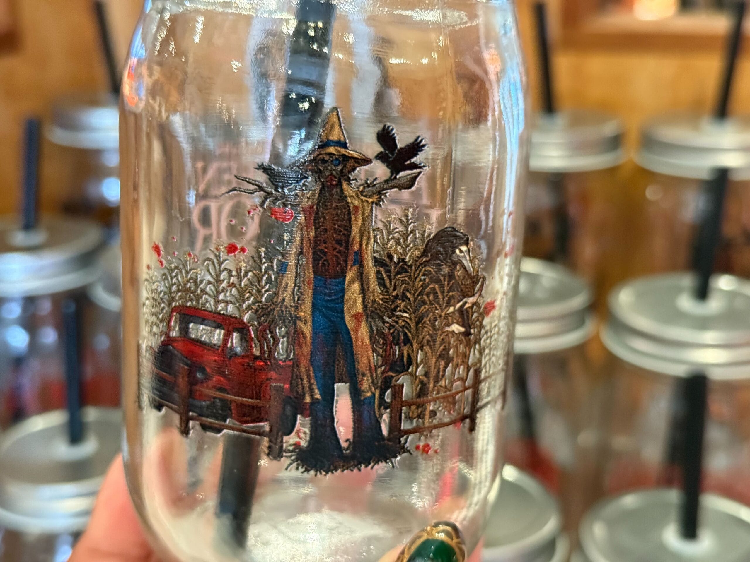 A hand holds a mason jar with a straw lid featuring the Scarecrow from the Scarecrow: The Reaping maze at Universal Studios Hollywood Halloween Horror Nights.