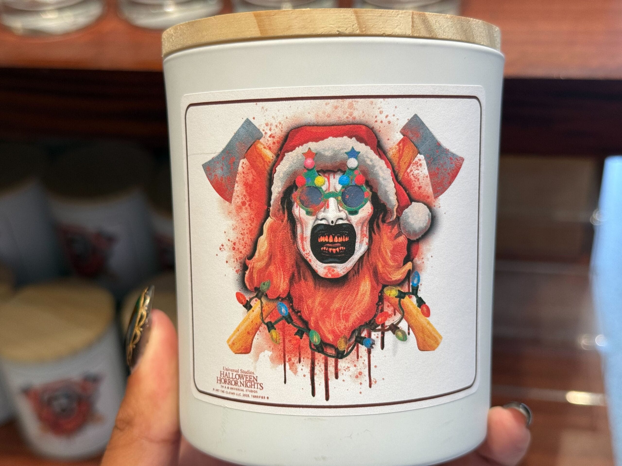 A 2025 Halloween Horror Nights lidded candle jar with Art the Clown in Christmas tree glasses and a Santa hat in front of bloody crossed axes.