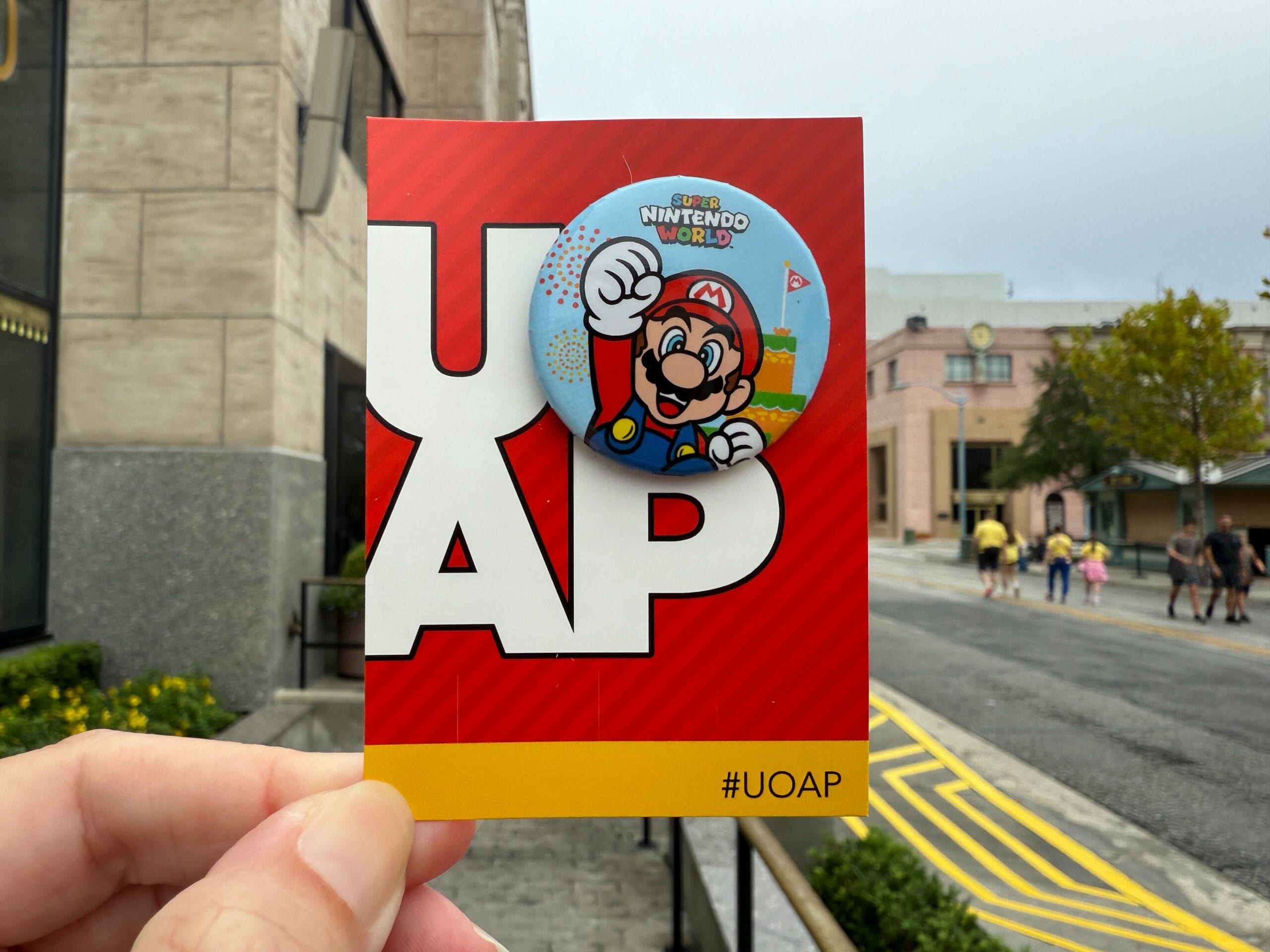 A hand displays a Super Nintendo World Mario pin card outdoors near Universal Orlando, not far from Disney castles like Cinderella's.
