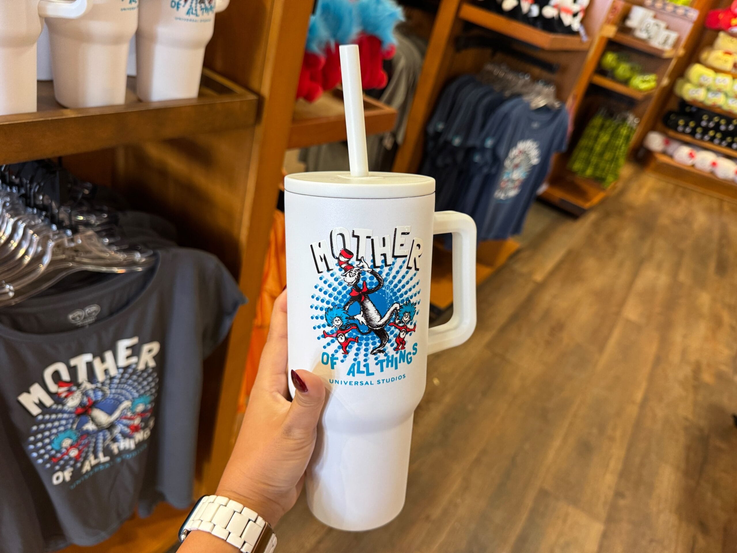 A hand holds a "Mother of All Things" mug with Dr. Seuss graphics in a gift shop, reminiscent of whimsical Disney park souvenirs.