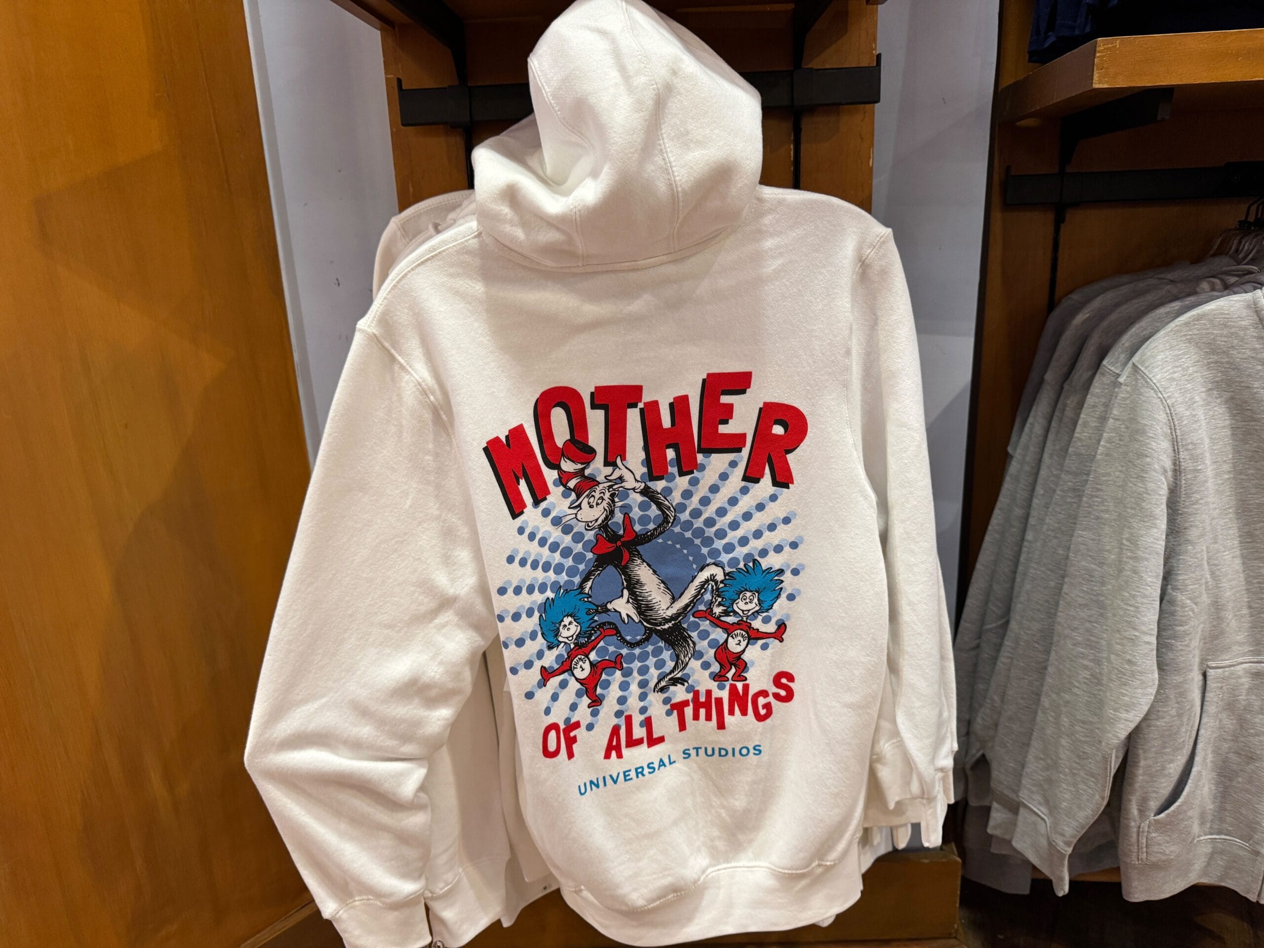 White hoodie with "Mother of All Things" and a cartoon cat riding Thing 1 and Thing 2, seen at Universal Studios—not a Disney castle.