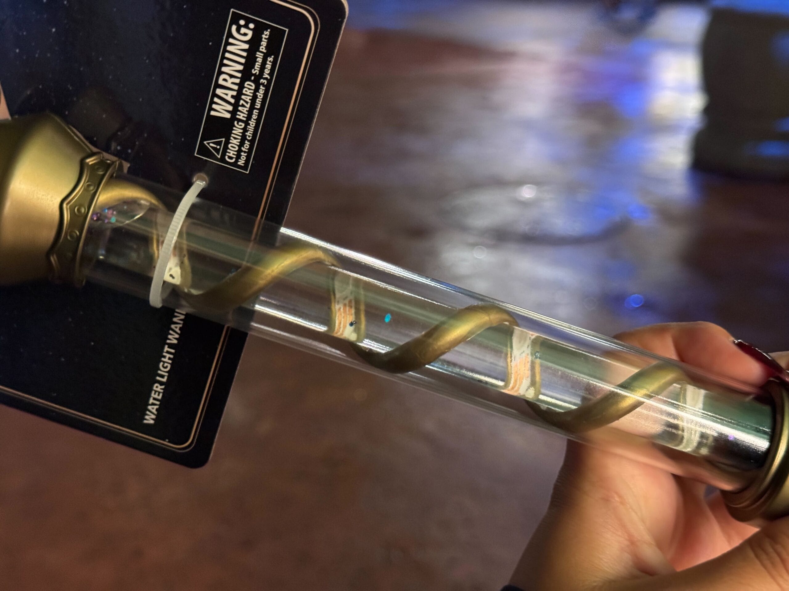 A hand holds a Celestial-Themed Water Light Wand with a golden spiral, under warm lighting in Epic Universe.