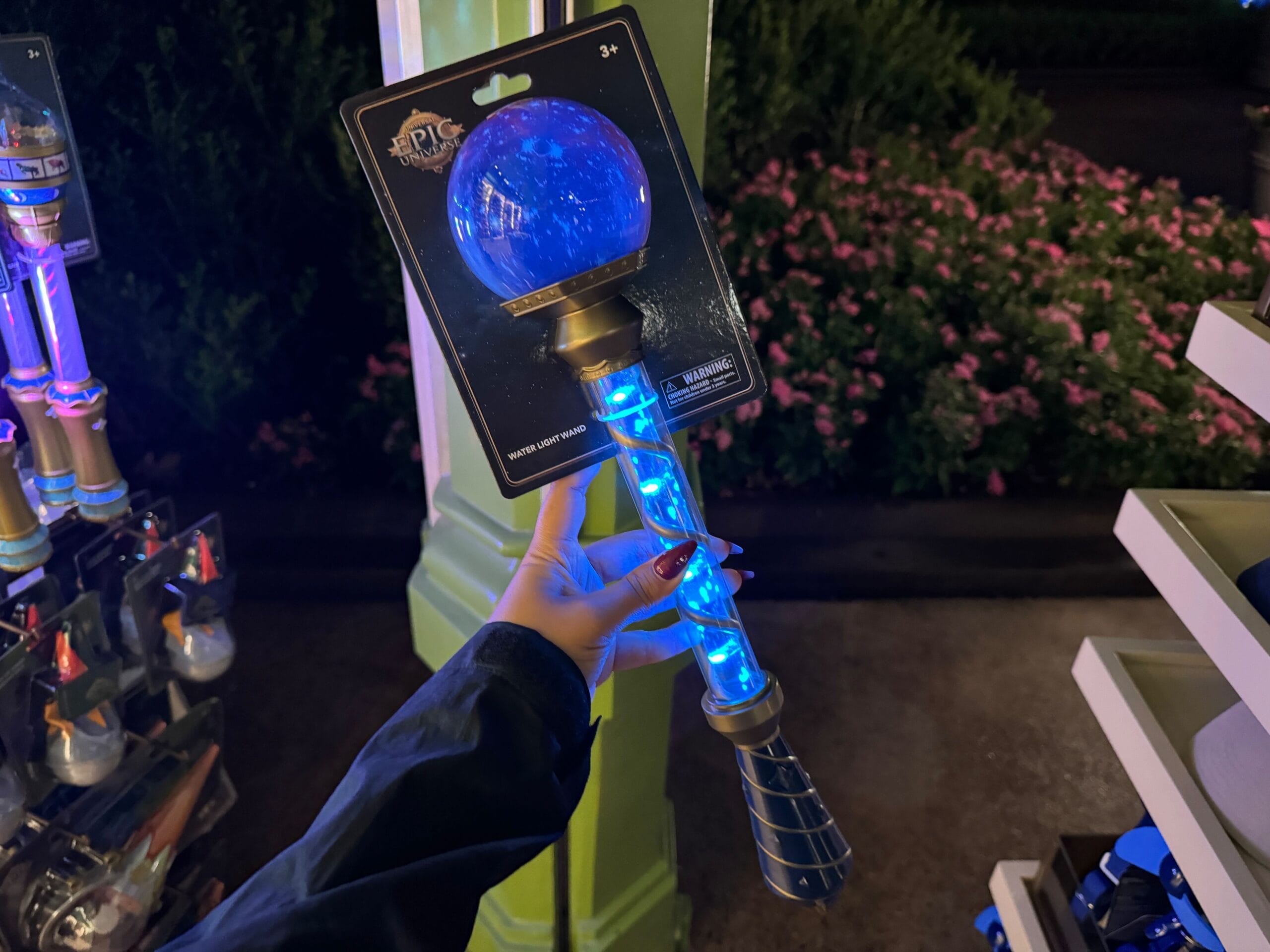 A hand holds a glowing Celestial-Themed Water Light Wand in packaging, with Epic Universe flowers and shelves behind.