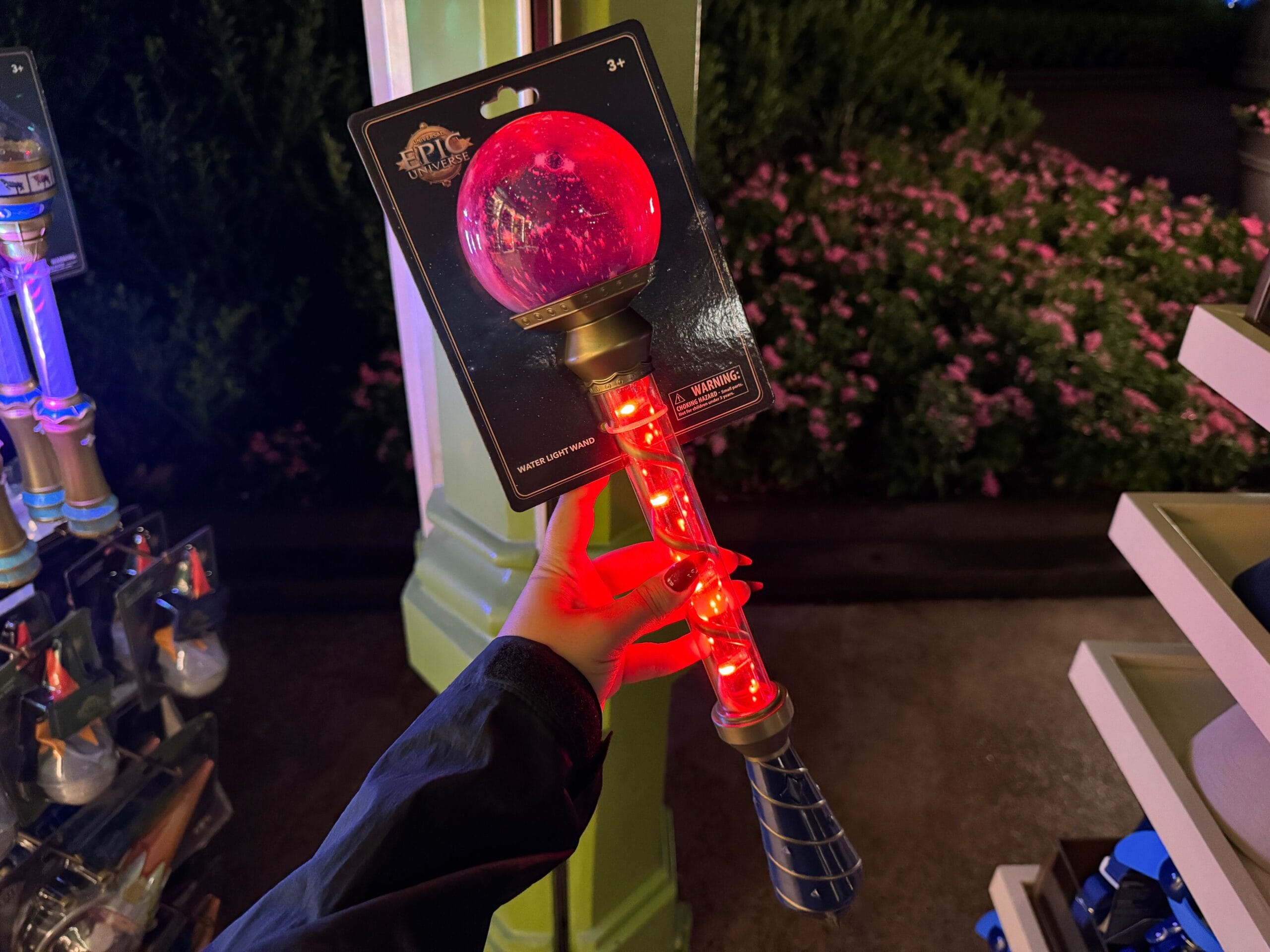 A hand holds a Celestial-Themed Water Light Wand with a glowing red orb in an Epic Universe outdoor shop at night.