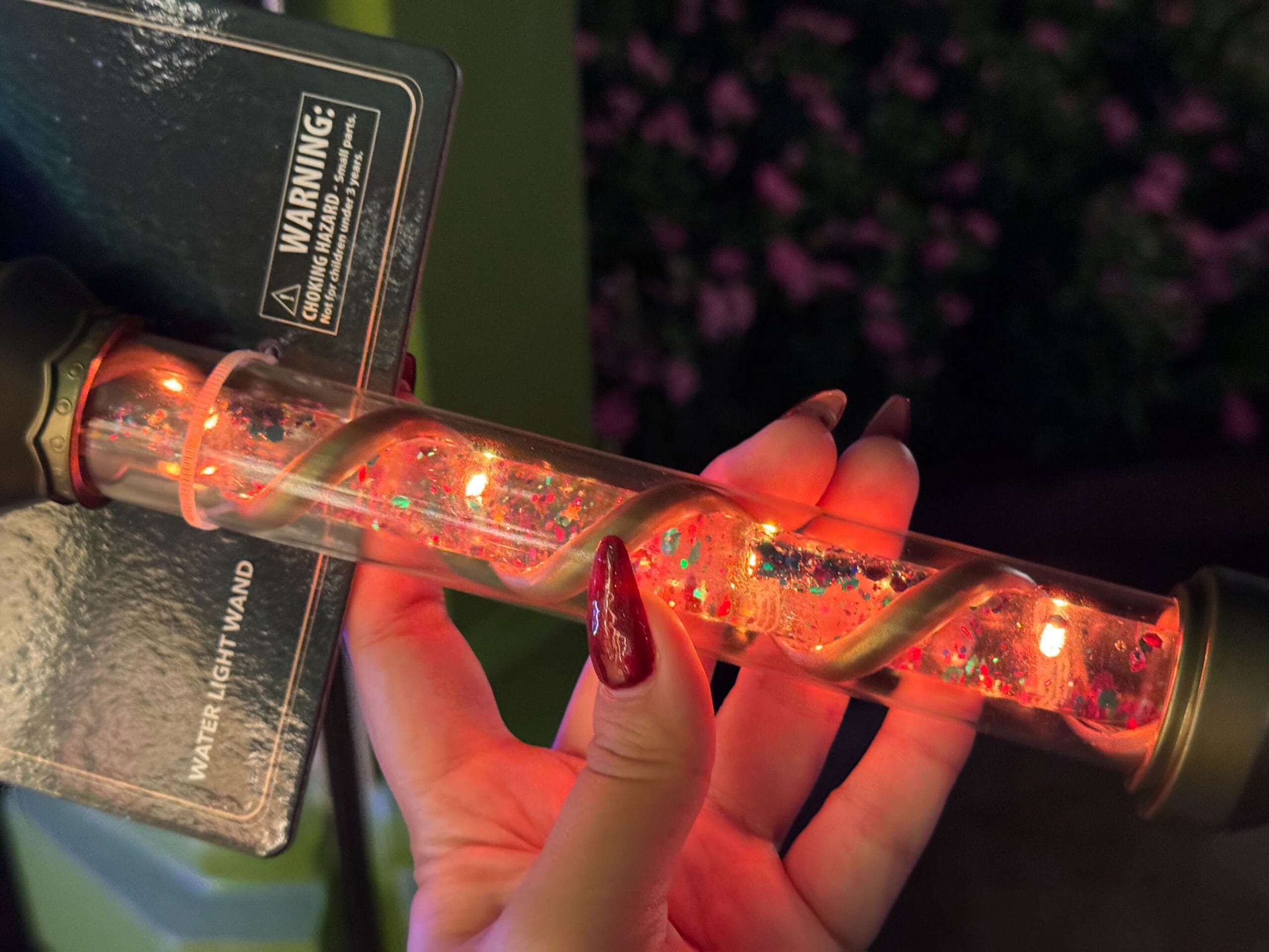 A hand with red nails holds an Epic Universe celestial-themed water light wand, glowing with sparkling orange-red lights.