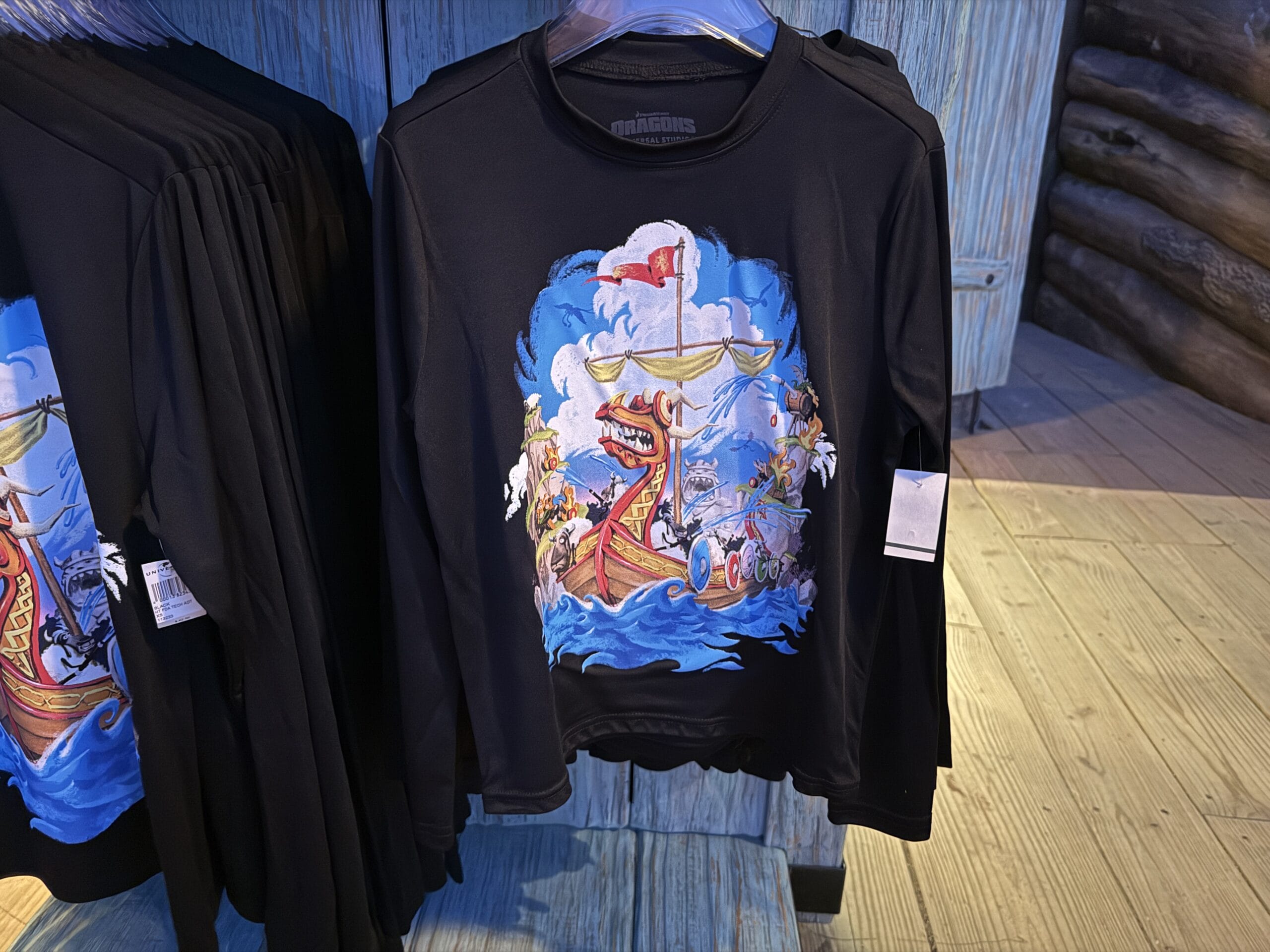 A black long-sleeve Spit Fyre shirt with a colorful cartoon Viking ship, displayed on a kiosk rack like Disney park merch.