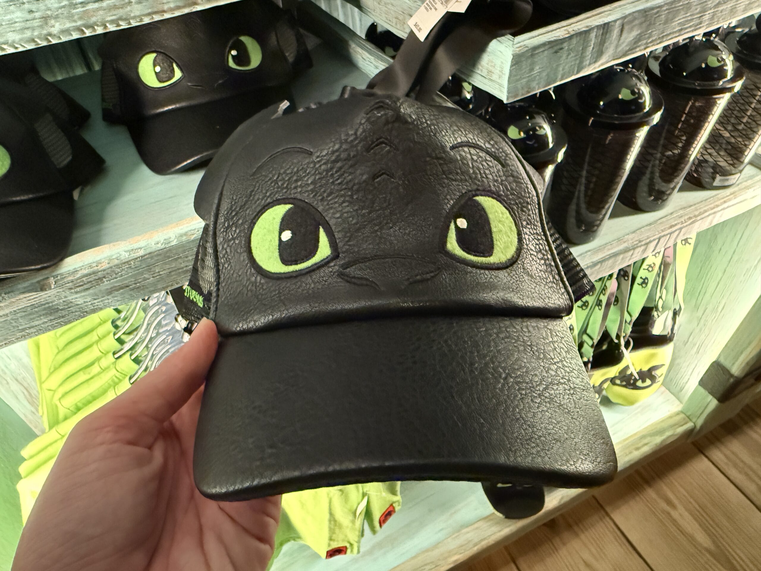 A hand holds a Toothless merch cap from How to Train Your Dragon, displayed at a store kiosk, much like Disney park shops.