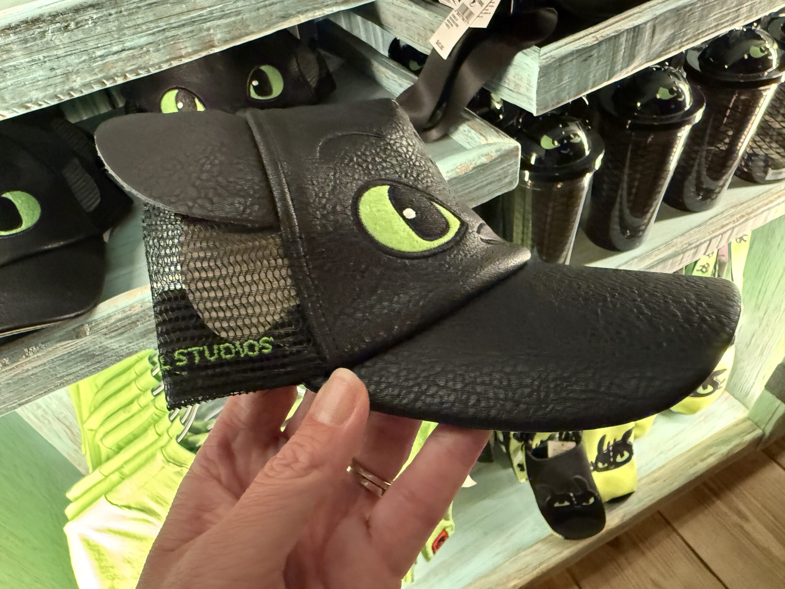 A hand displays a Toothless dragon cap with green eyes and pointy ears, sold at the Spit Fyre kiosk—no Disney castle in view.